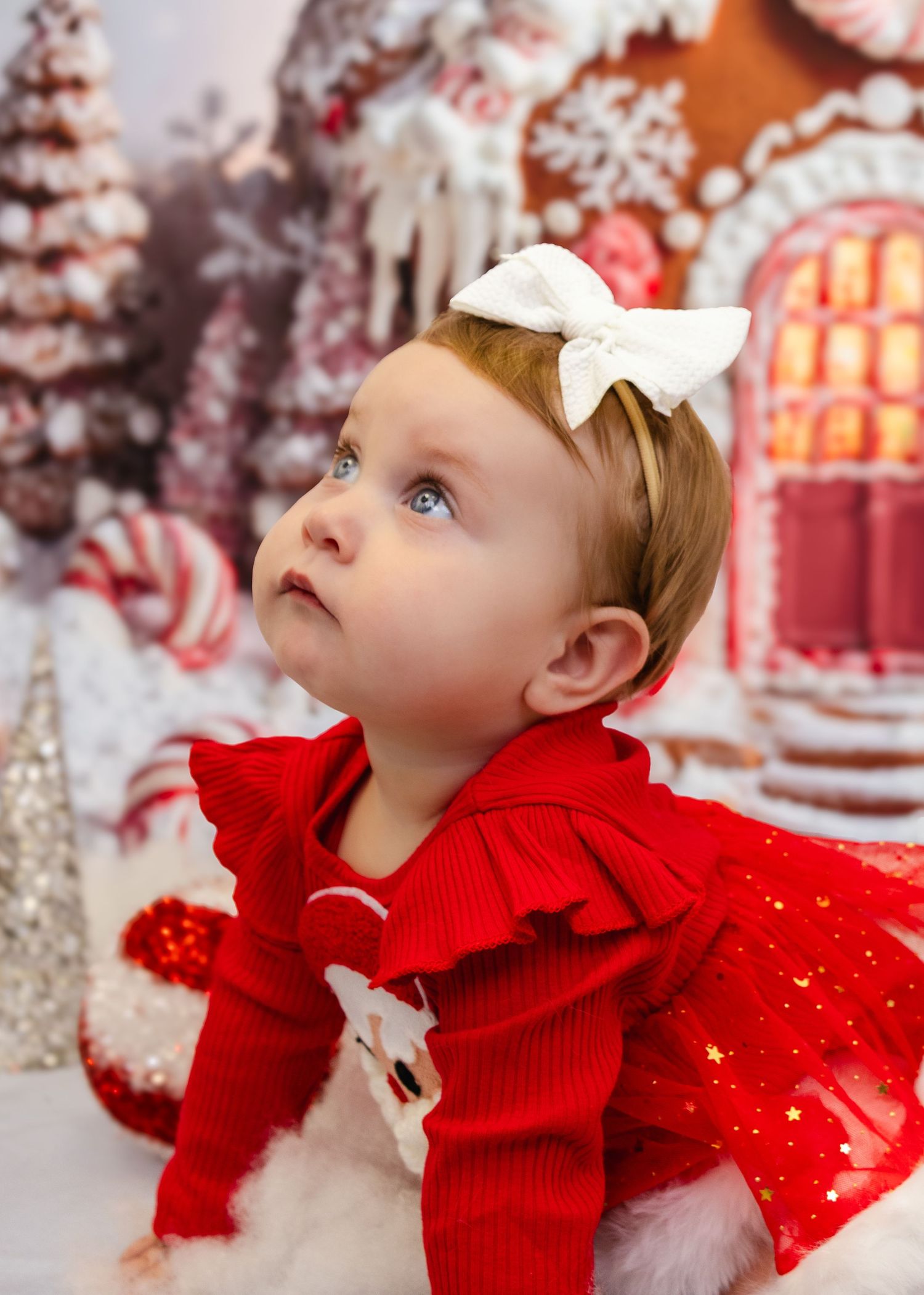 Baby girl in red Christmas outfit with white bow gazes upward before a festive gingerbread house backdrop.