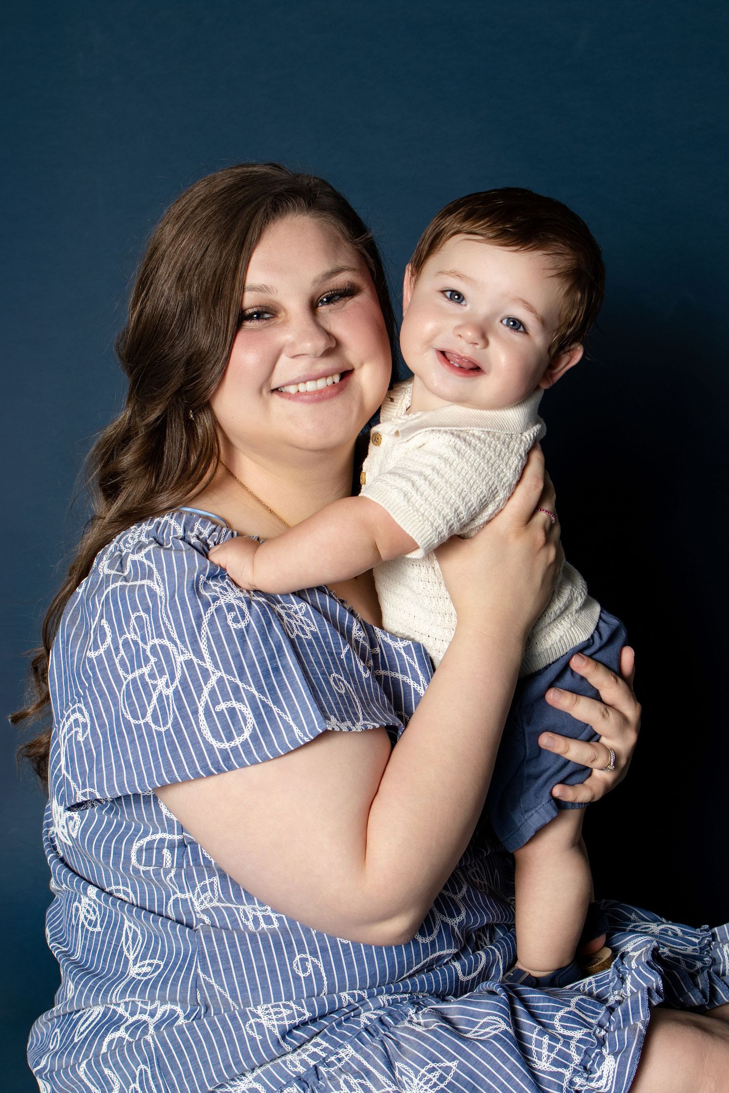 A smiling young woman in a blue dress holds a happy baby boy against a dark blue studio background.