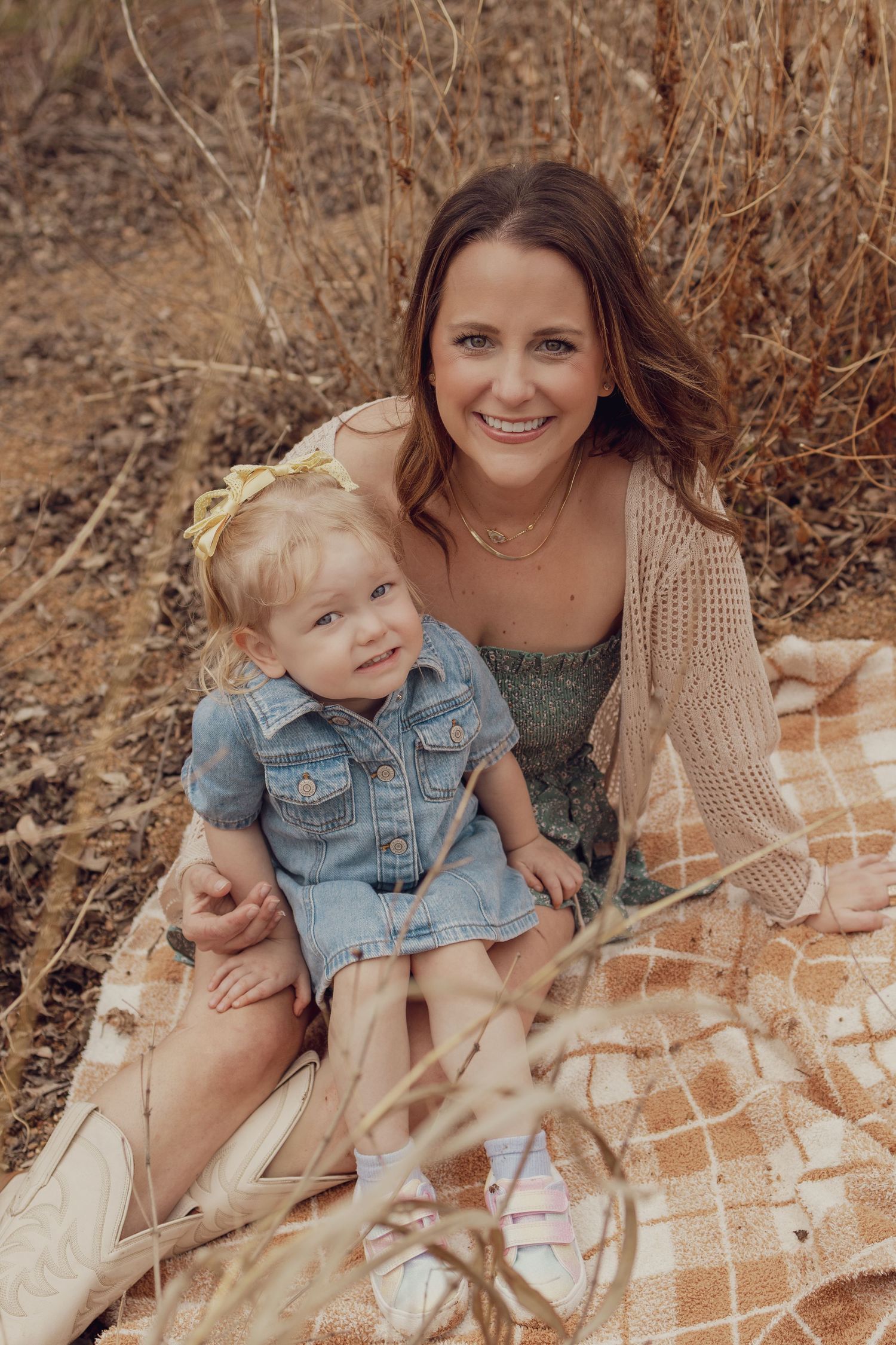 Smiling mother sitting on plaid blanket outdoors holding her toddler daughter in a denim dress.