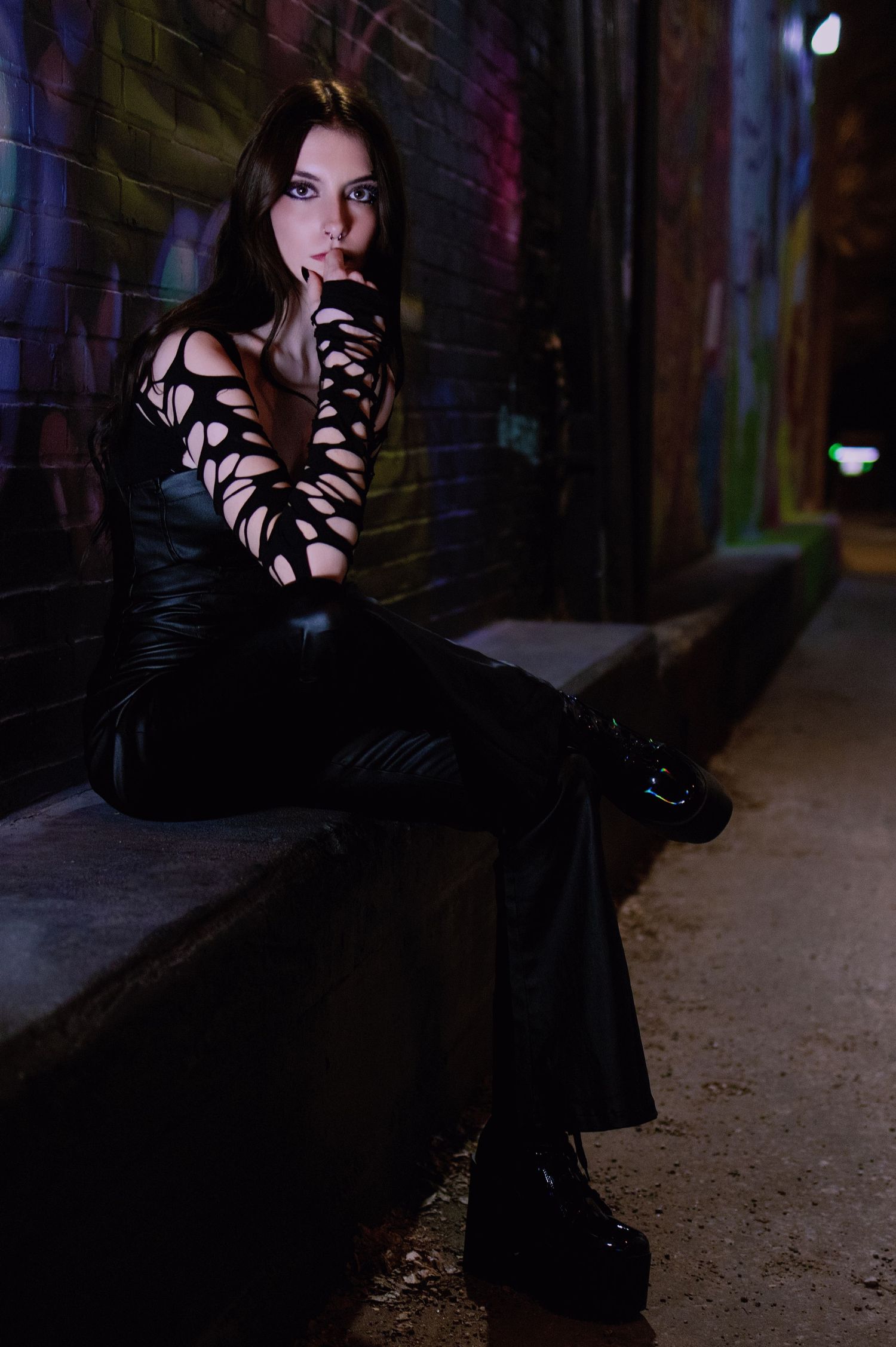 Woman in black leather outfit with cutout sleeves sits in dark graffiti alley at night.