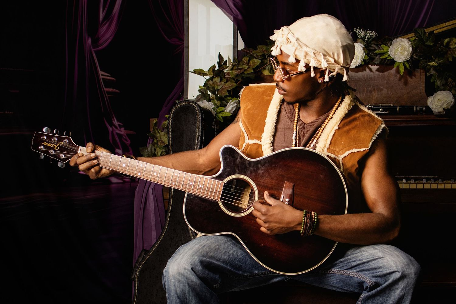 A musician plays acoustic guitar wearing a fur vest and decorative head wrap in a dramatic studio setting.