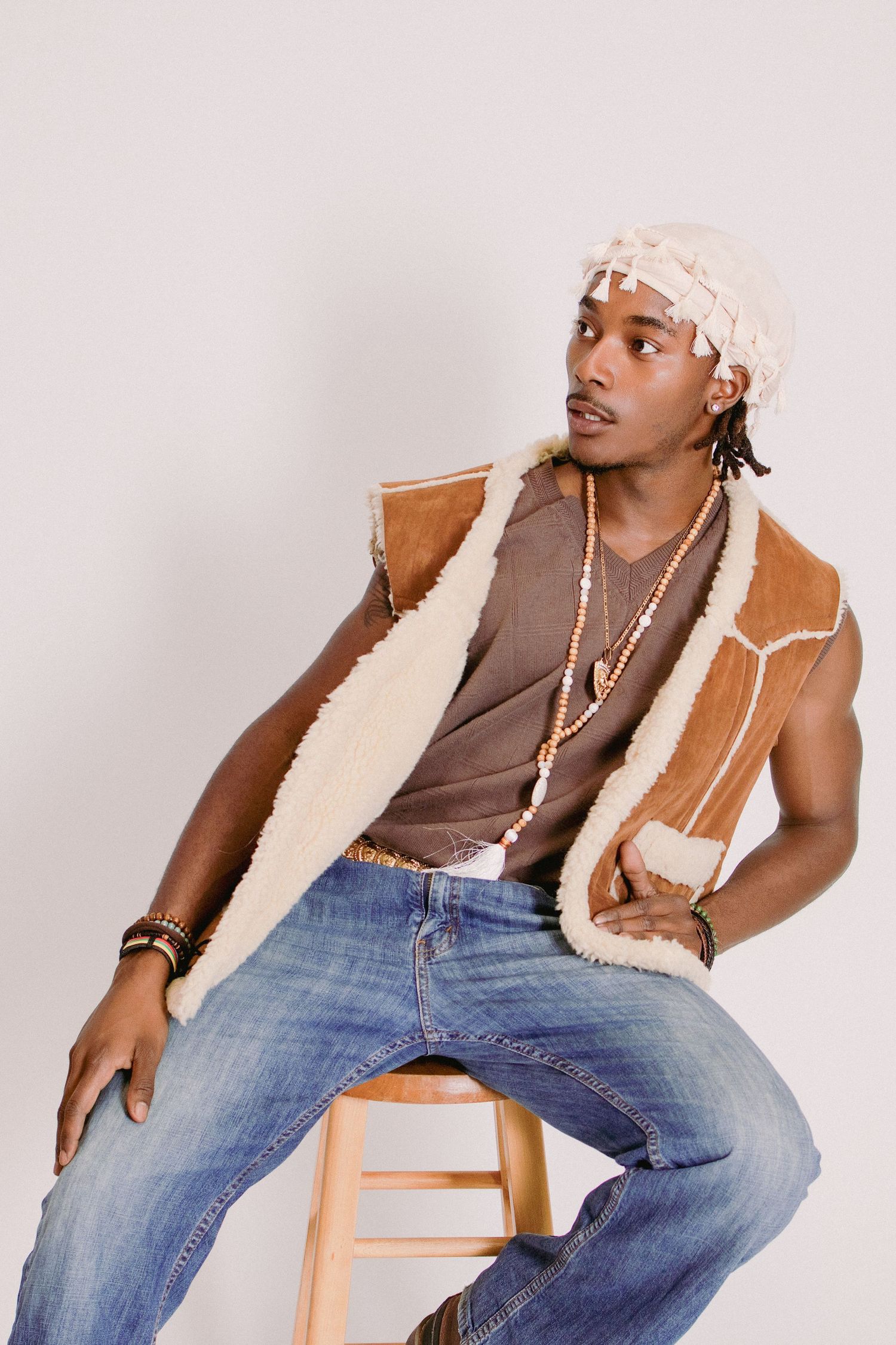 Young Black man sitting on stool wearing shearling vest, jeans, beaded necklace, and white durag in studio.
