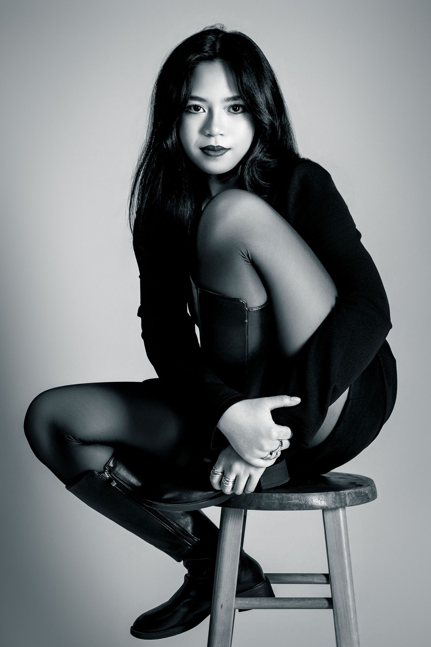 Black and white studio portrait of woman in black outfit sitting on wooden stool.