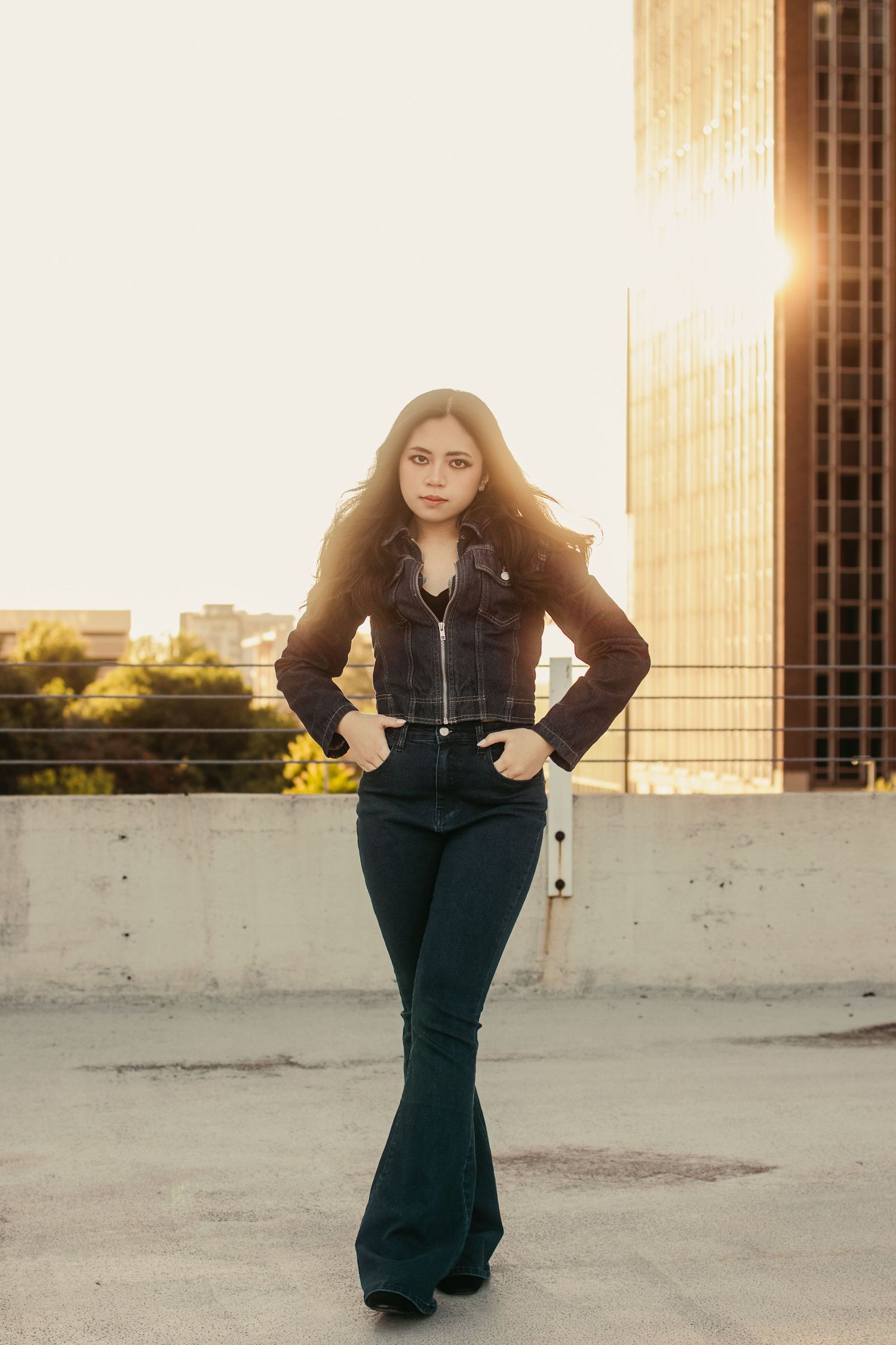Young woman in black jacket and flare jeans posing on rooftop at sunset with urban skyline background.