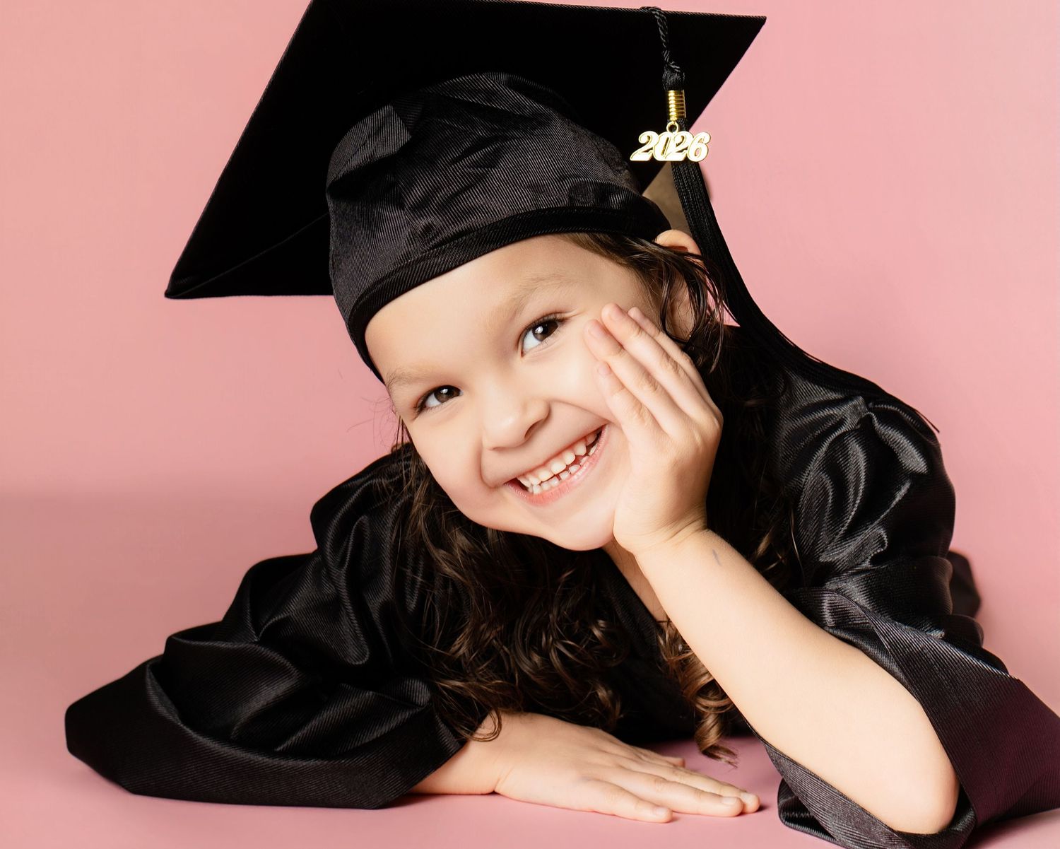 Smiling young girl in black graduation cap and gown posing against a pink background.