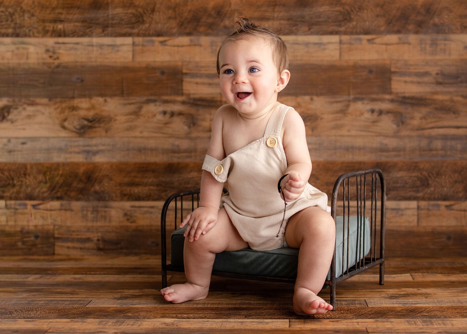 Smiling baby in beige romper sitting on mini metal bed against rustic wood plank background.
