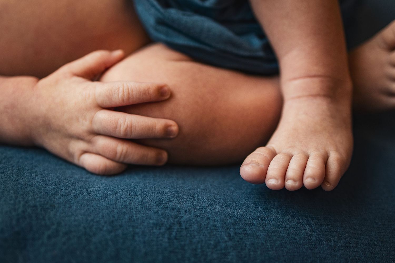 Close-up of an adult hand gently holding a baby's chubby legs and tiny toes on a blue surface.