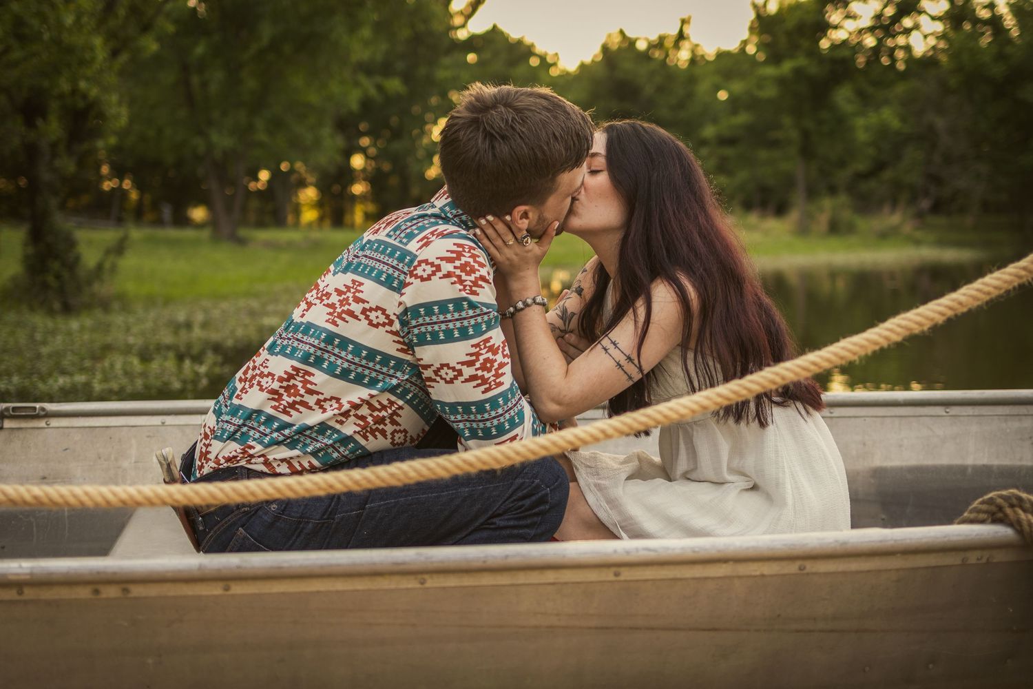 Young couple kissing tenderly in a wooden rowboat surrounded by lush greenery at golden hour.