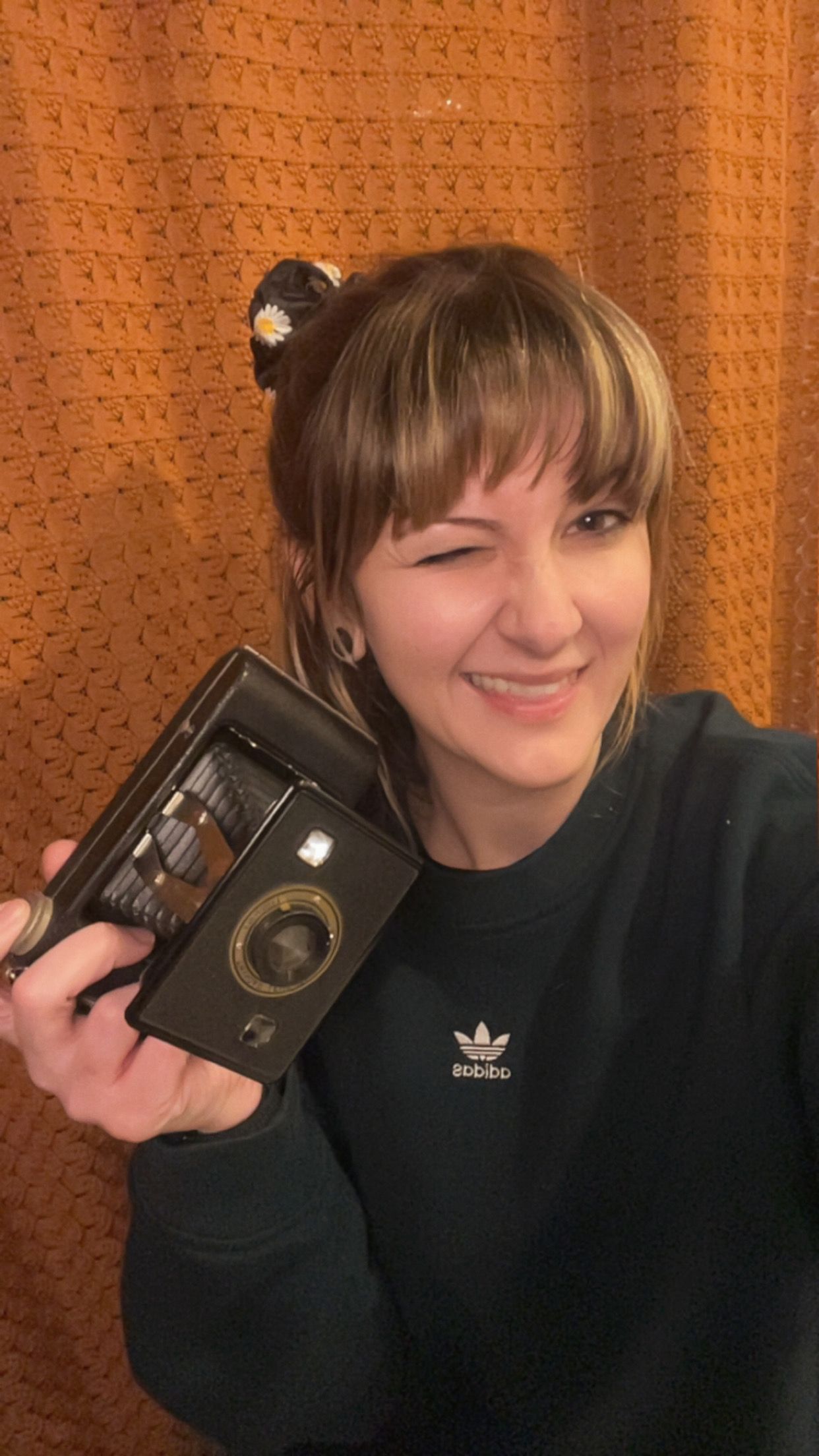 A smiling young woman with bangs holds a vintage black camera, wearing an Adidas sweatshirt against an orange curtain.