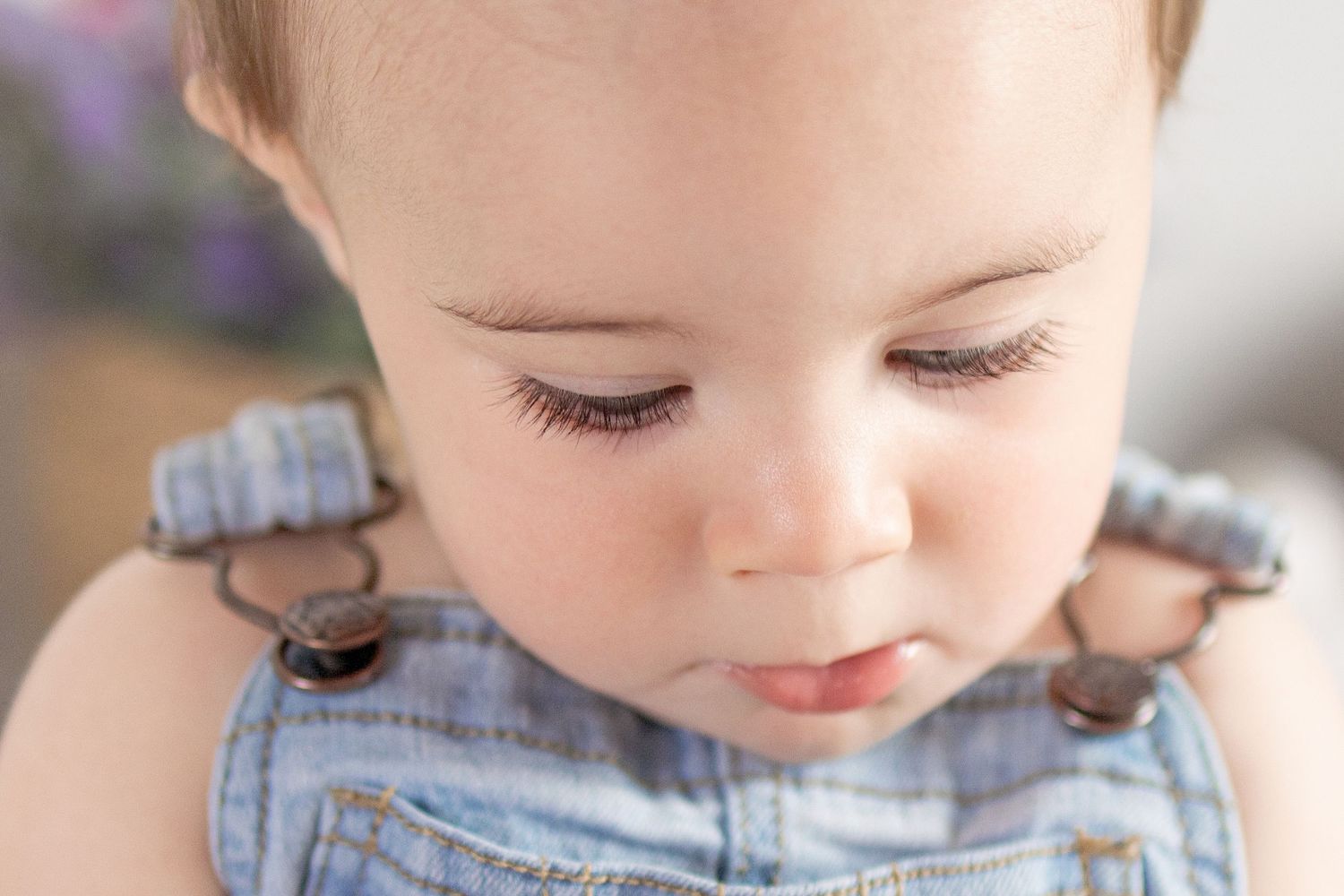 Close-up portrait of a cute baby with long eyelashes wearing denim overalls, looking down with a curious expression.