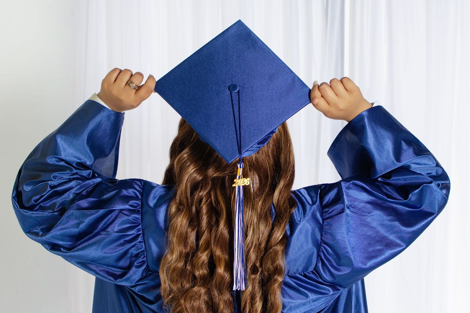 Back view of a student in blue graduation cap and gown with long brown wavy hair.