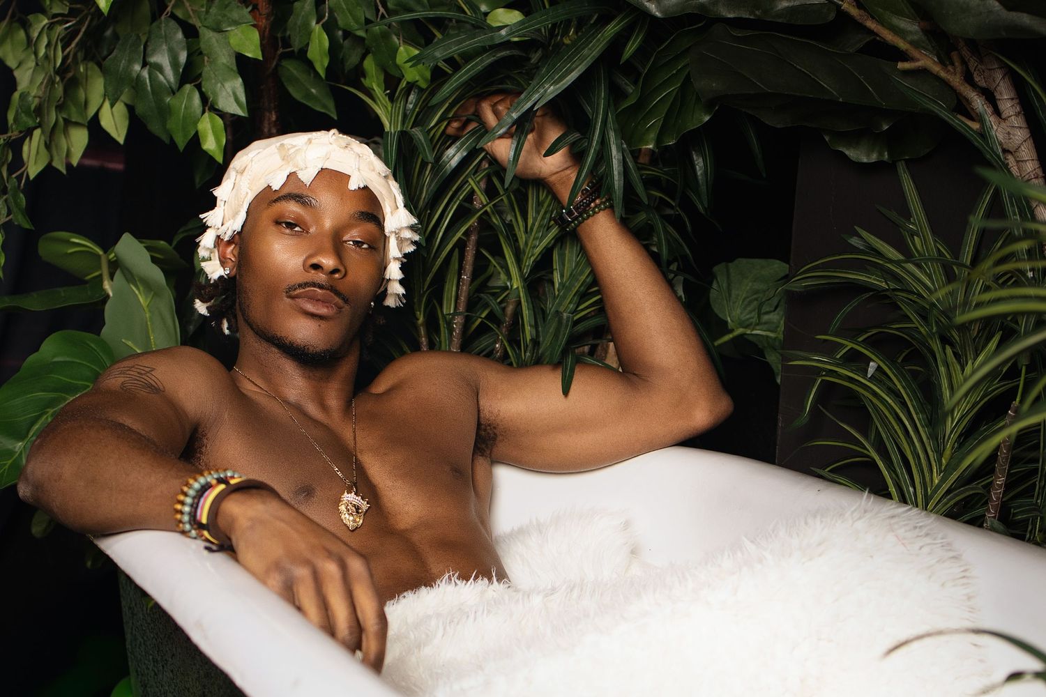 Shirtless man relaxing in a white bathtub surrounded by lush tropical plants, wearing gold jewelry and a white headband.