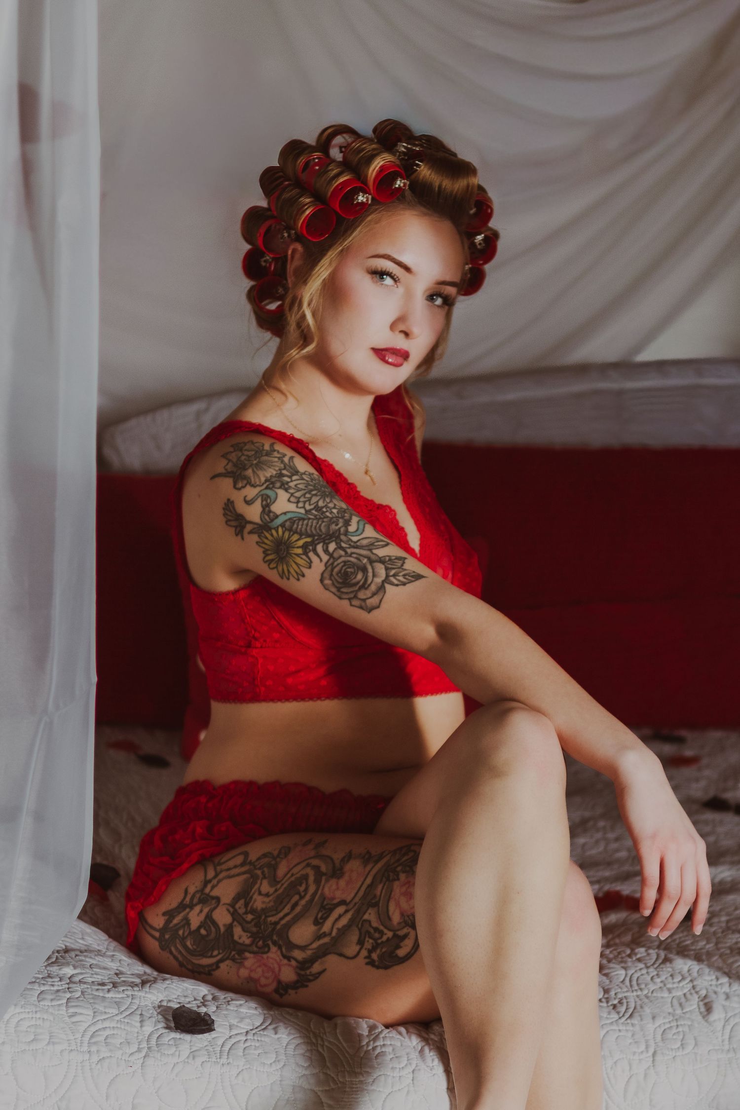 Tattooed blonde woman in red lace lingerie and hair curlers sitting on a bed with white drapes.
