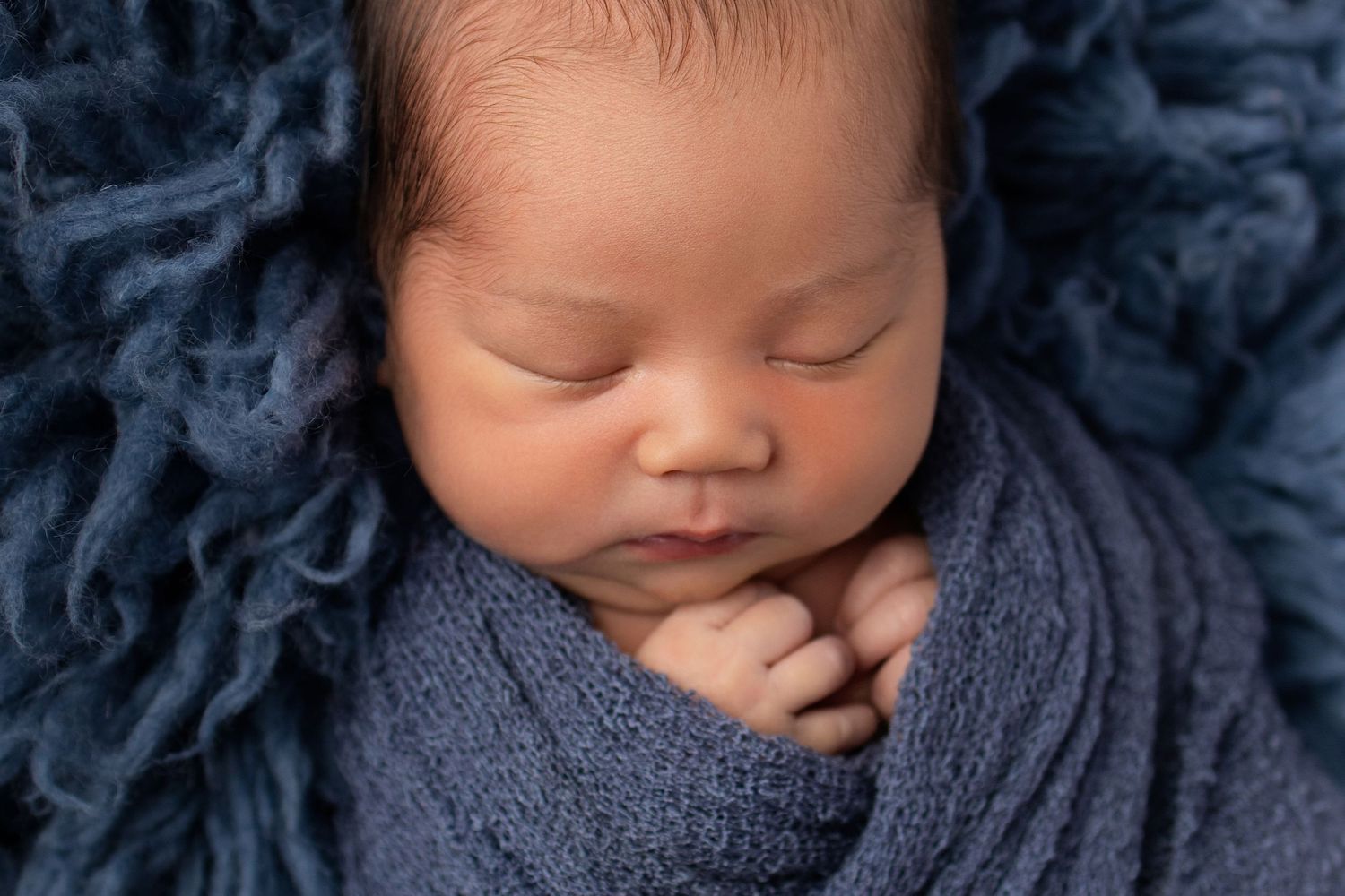 Sleeping newborn baby wrapped in a blue knit blanket on a fluffy blue rug for a professional photo.