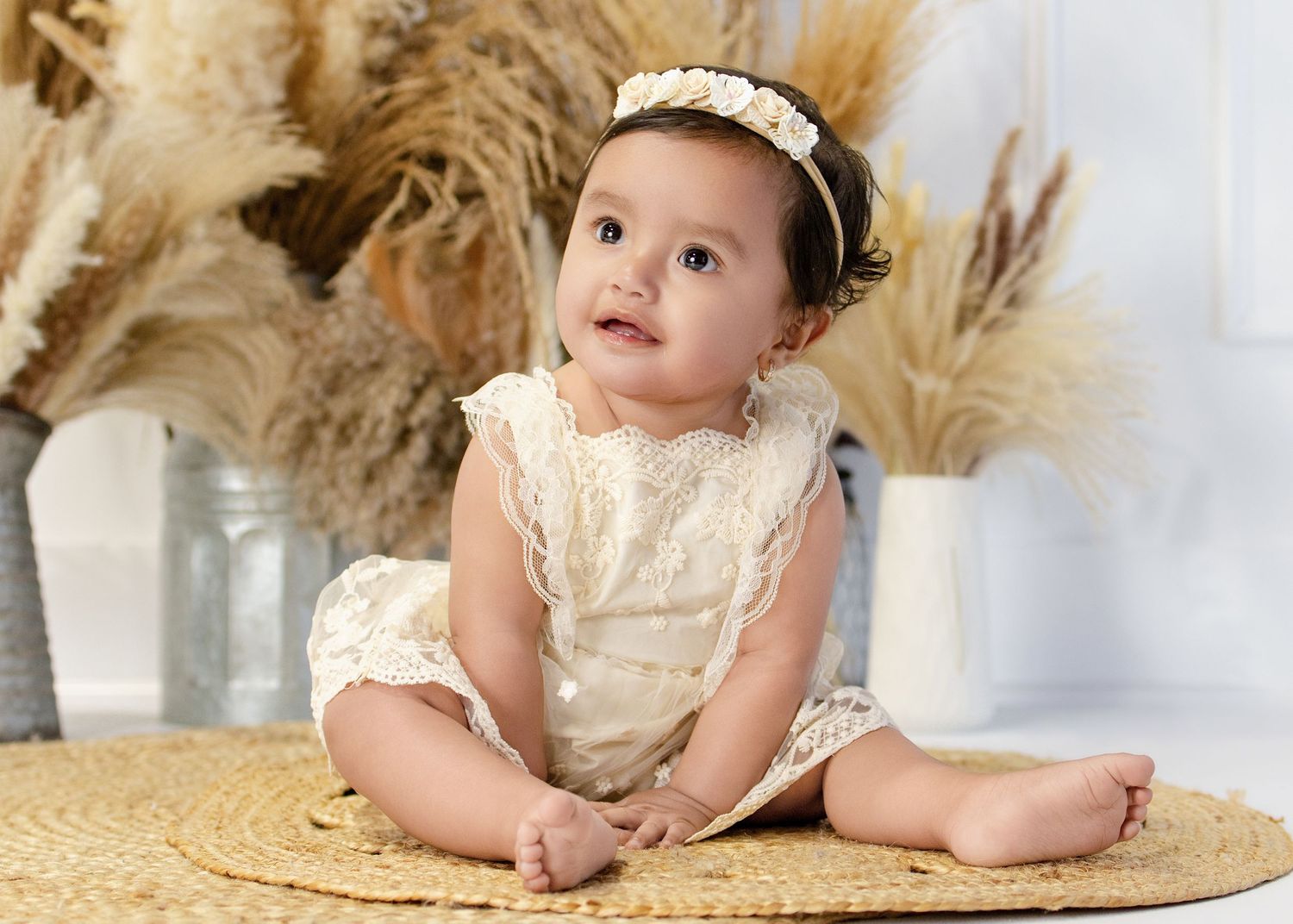 Adorable baby girl in cream lace romper and floral headband sits on woven mat with dried pampas grass decor.