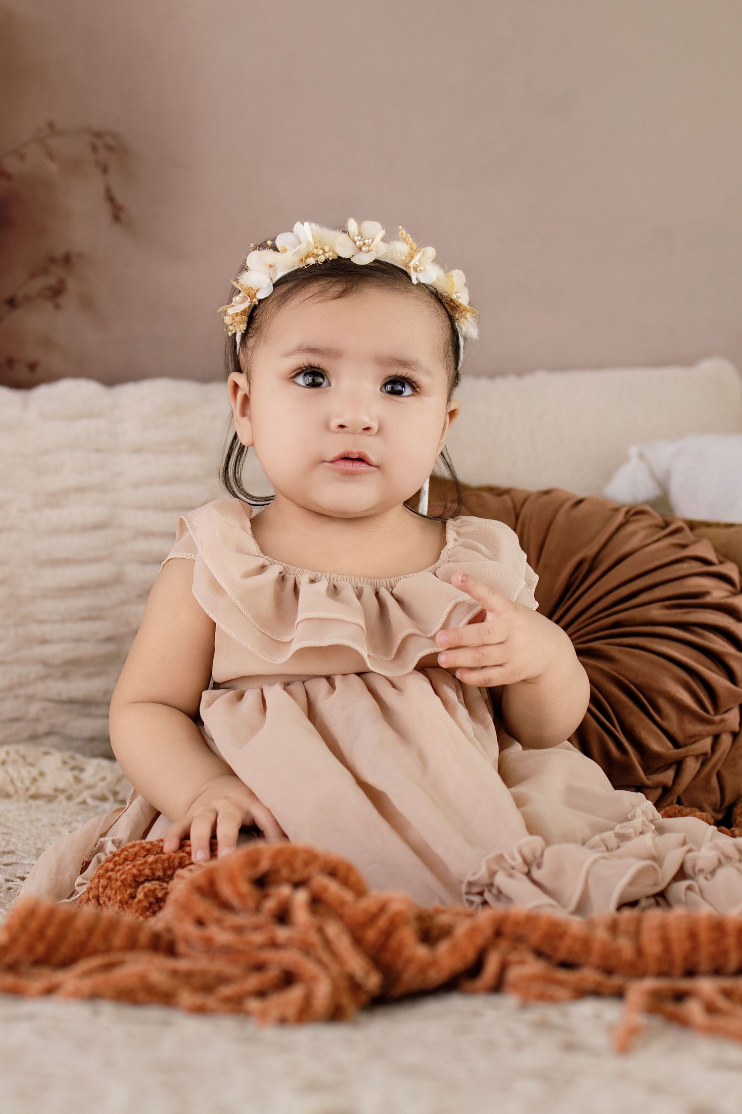 Adorable baby girl in a blush ruffle dress and floral crown sits on a cozy blanket for a portrait photo.