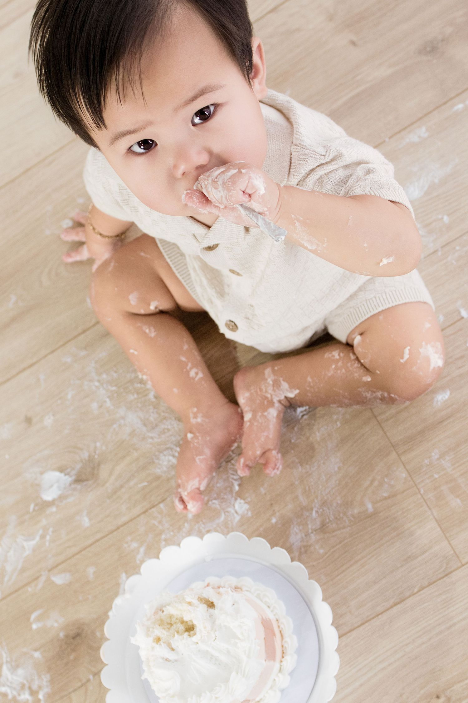 Overhead view of baby eating smashed white birthday cake off floor during first birthday celebration.