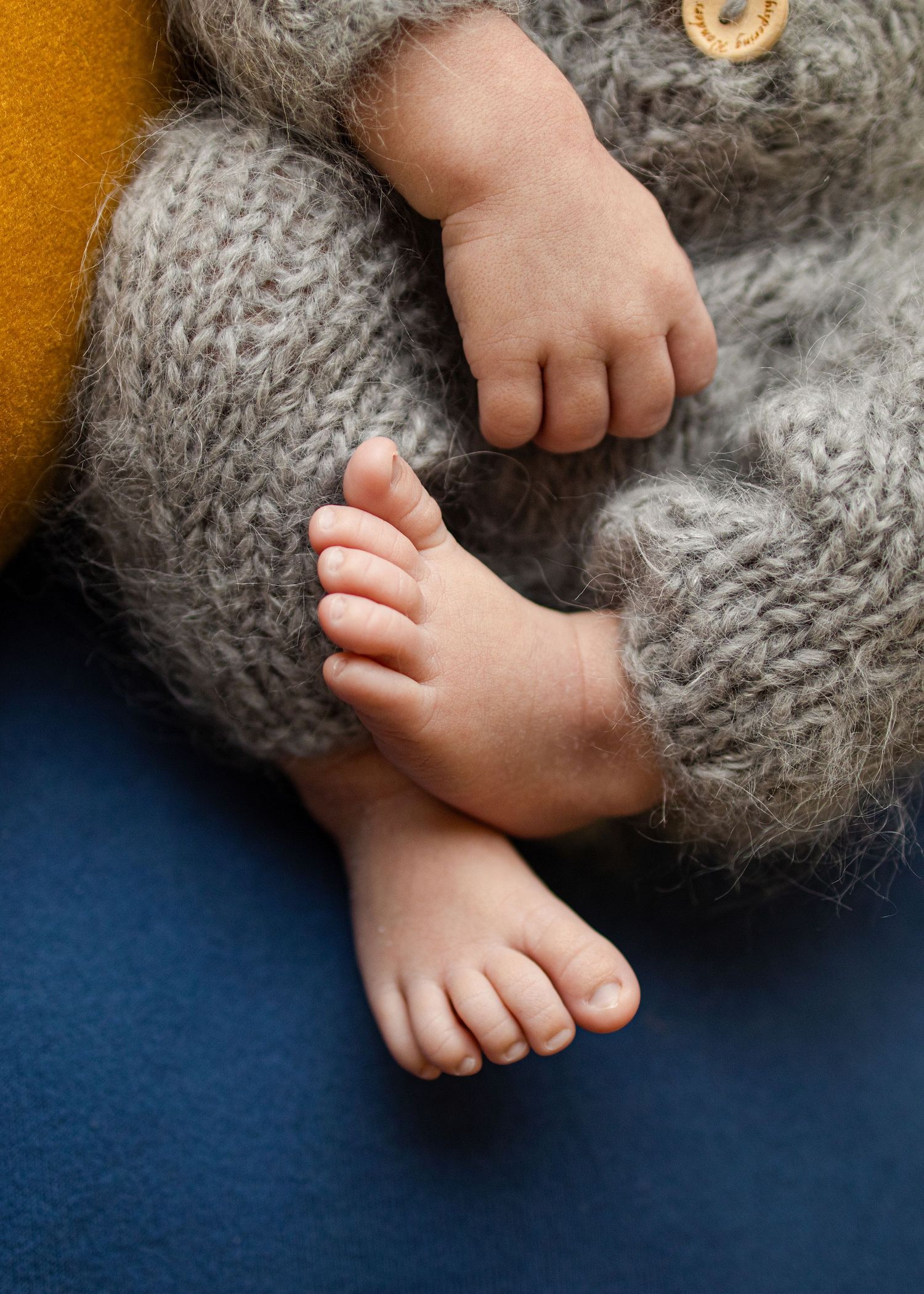 Close-up of a newborn baby's tiny feet and hand wrapped in a soft gray knit sweater on a blue surface.