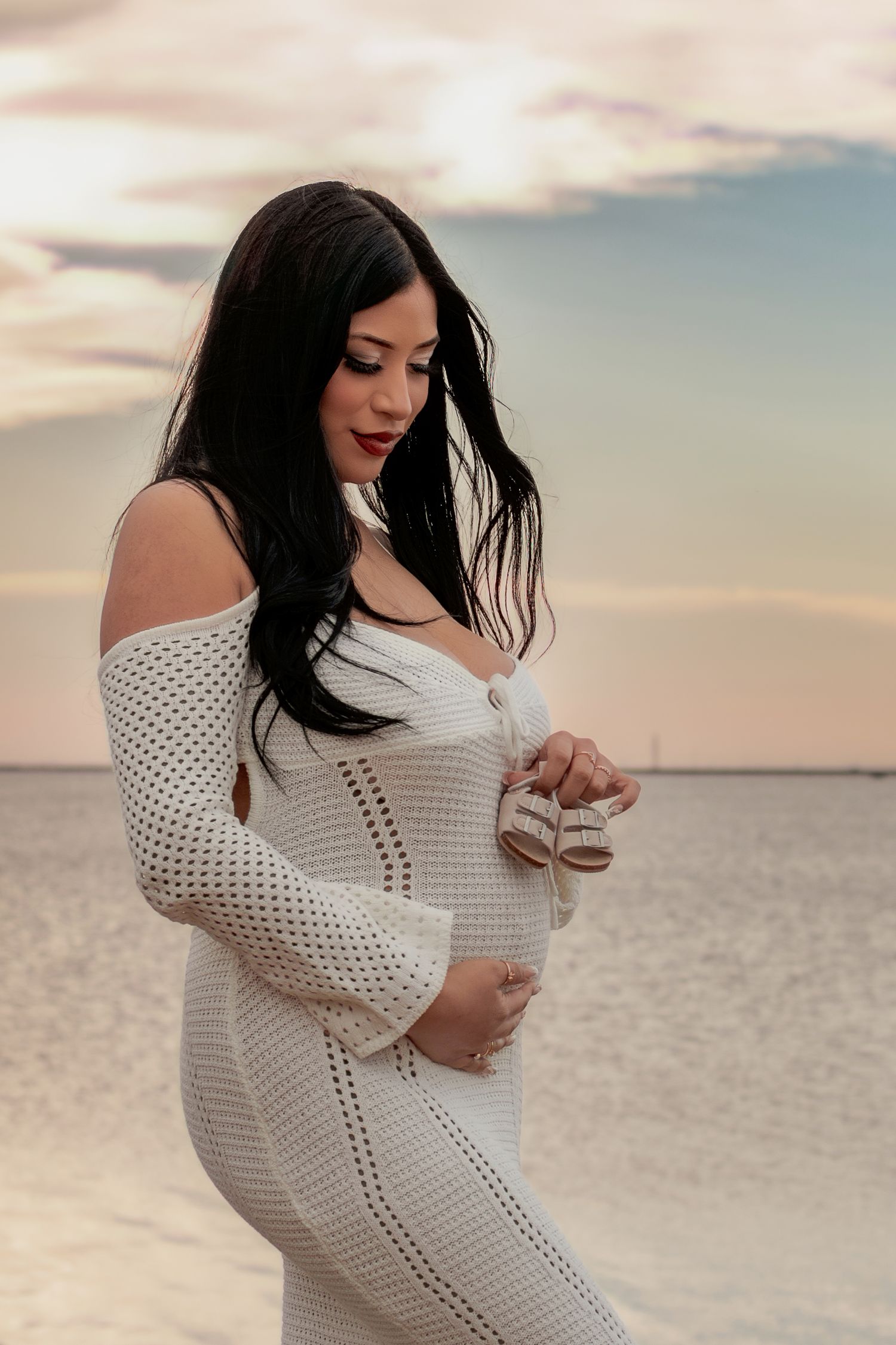 Pregnant woman in white knit dress holding baby shoes on beach at sunset.