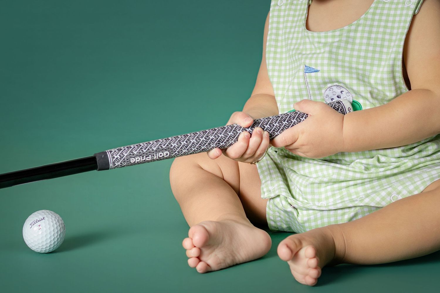 Baby in green outfit holding a golf club grip with a white golf ball nearby on a green background.