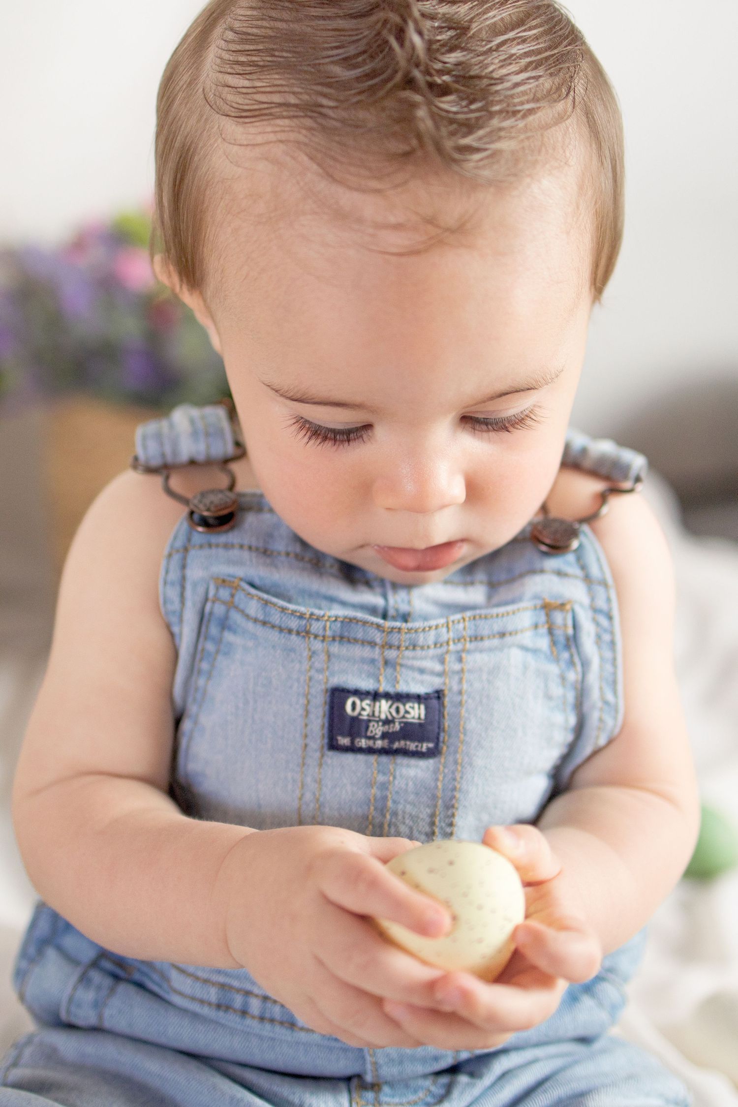 Adorable baby in OshKosh denim overalls carefully examining a small egg with curiosity.