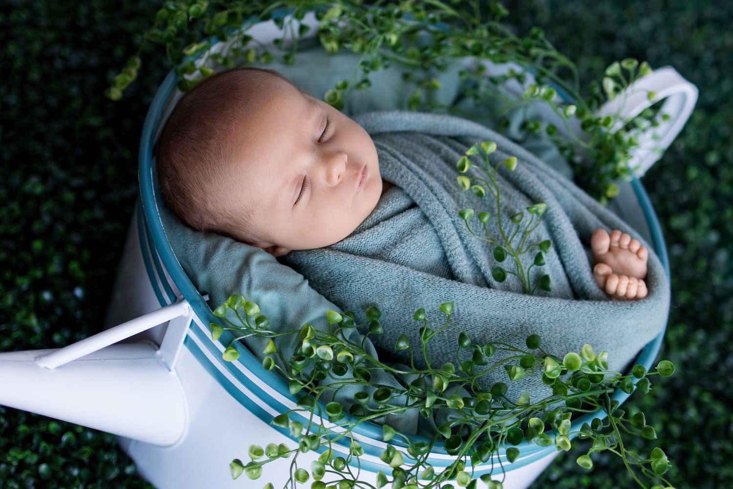 A baby rests peacefully in a blue wrap hammock surrounded by delicate green foliage and white flowers outdoors.