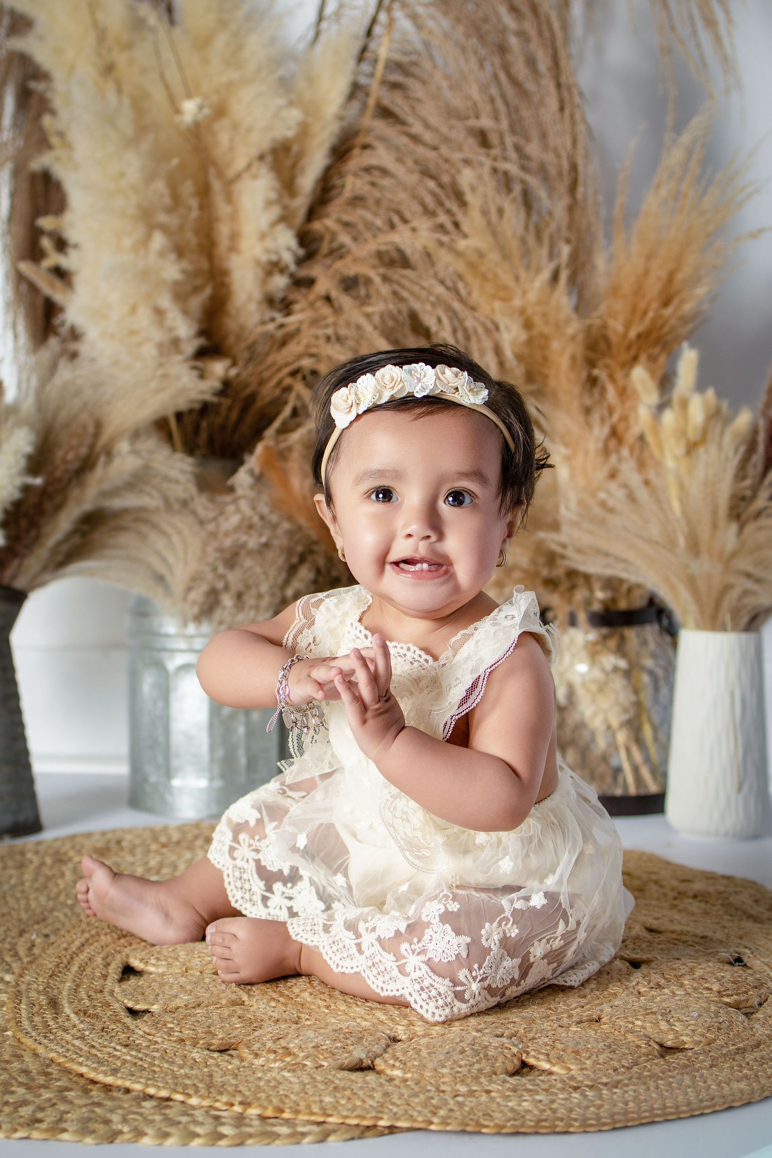 Smiling baby girl in cream lace romper and floral headband sitting on woven rug with dried pampas grass decor.