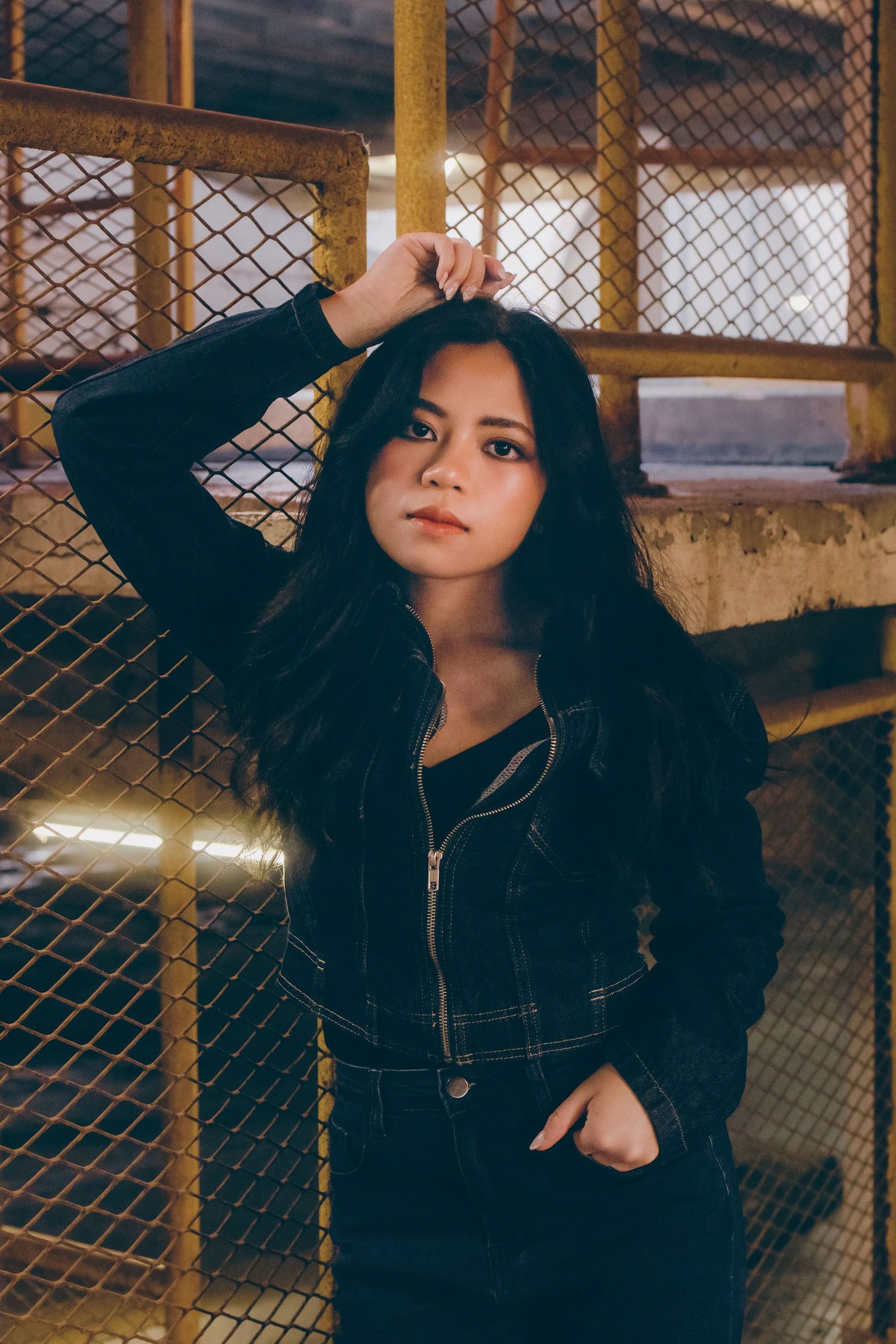 Young woman in black denim jacket poses confidently against chain-link fence in urban setting.