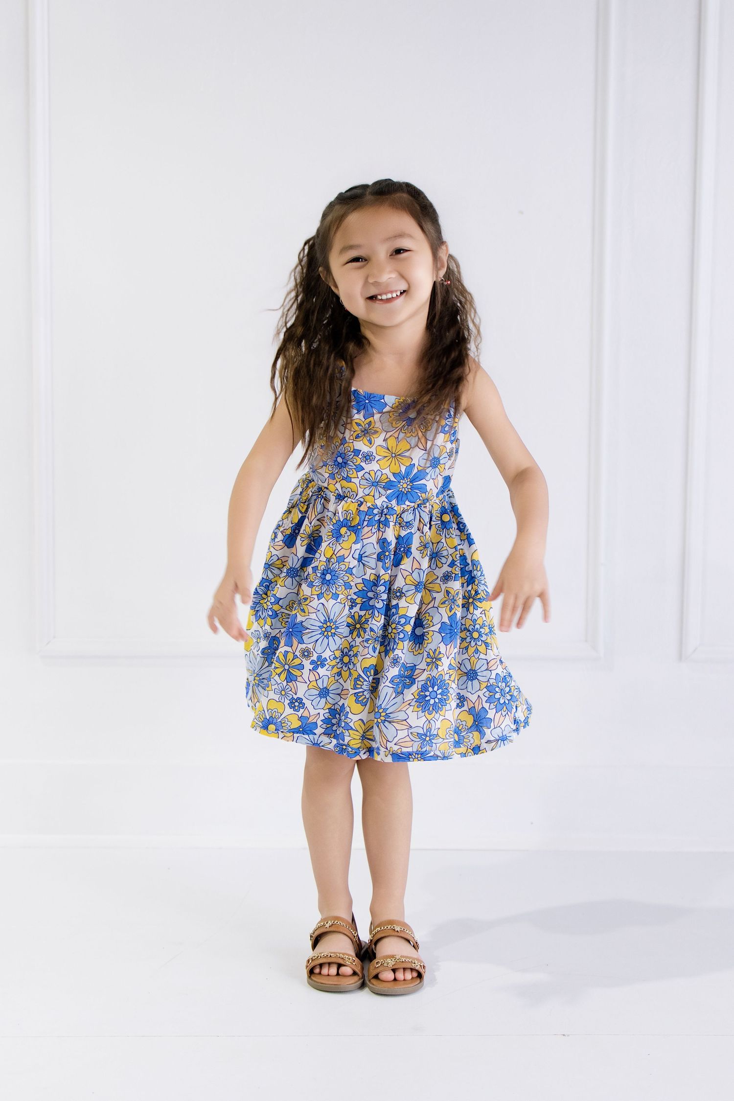 Young girl smiling in a blue floral sundress and sandals, posing playfully against a white background.