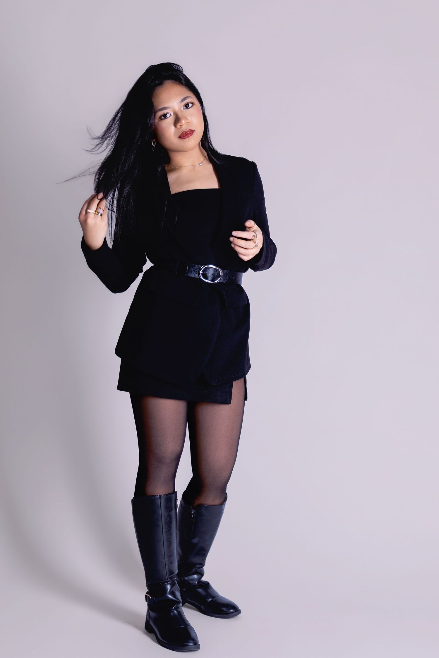 Woman in black blazer dress, belt, tights, and knee-high boots poses confidently against a light gray background.