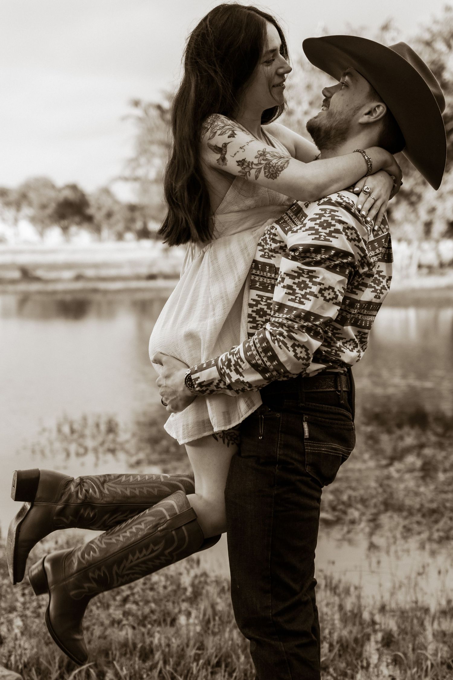Sepia-toned photo of a man in cowboy hat lifting a woman in cowboy boots near a pond.