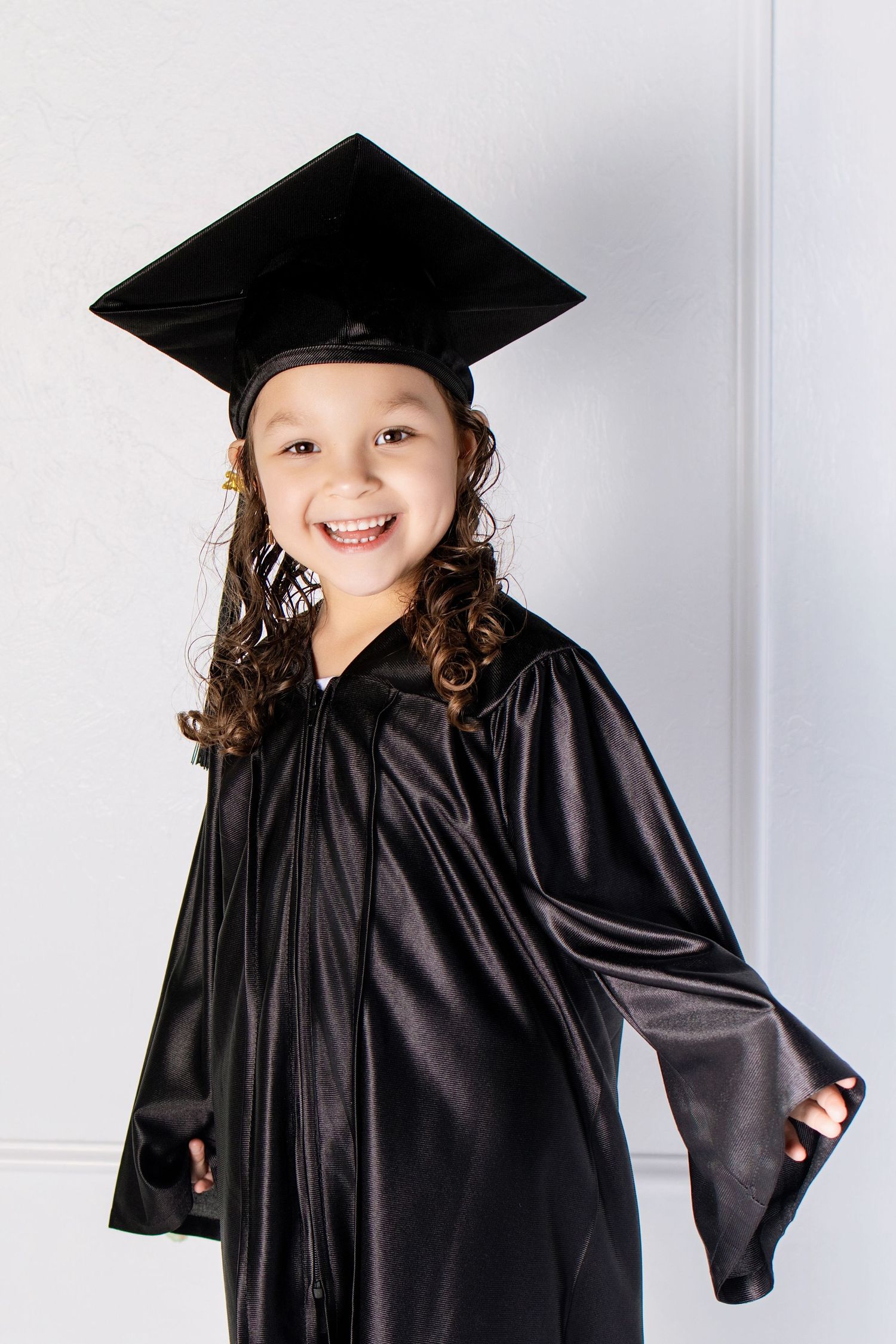 A smiling young girl wearing a black graduation cap and gown, posing proudly against a white wall.