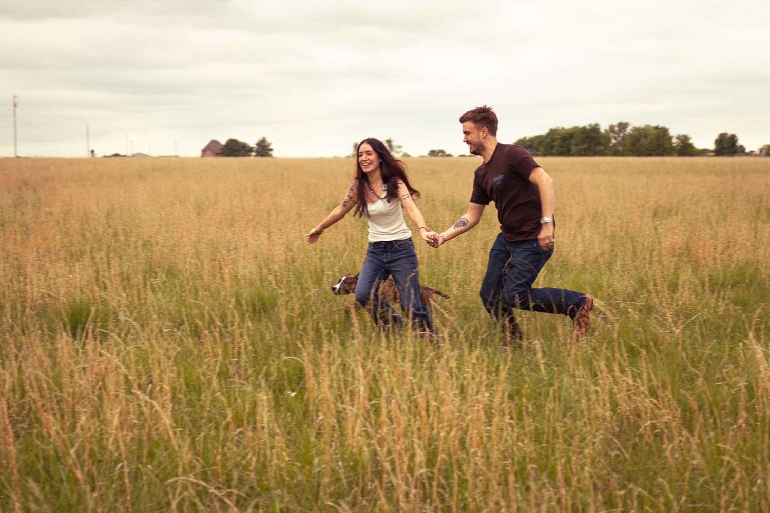 A joyful young couple runs hand-in-hand through a golden meadow field under a cloudy sky.