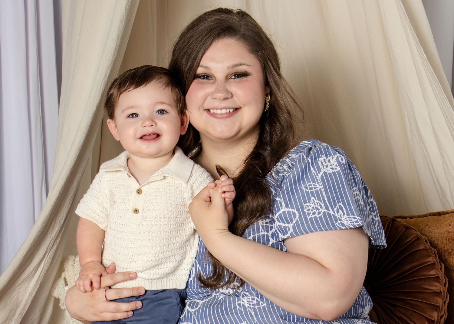 A smiling young woman and toddler boy pose together in a cozy tent backdrop for a professional portrait photo.