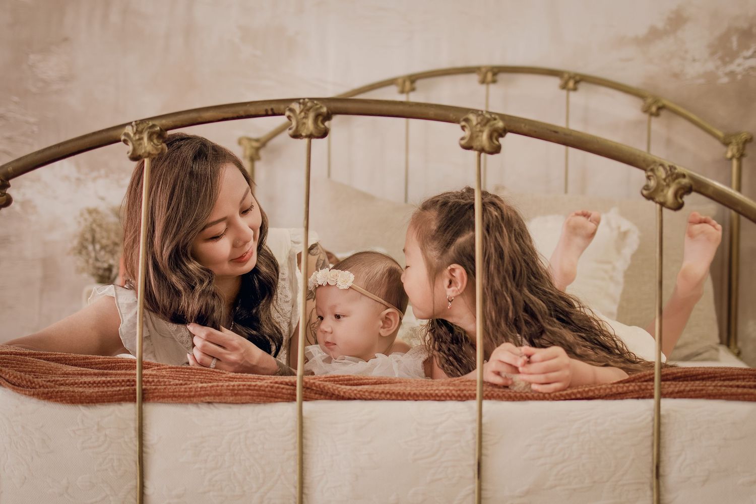 A mother and two young daughters share a tender moment lying on a vintage brass bed in a soft, dreamy setting.