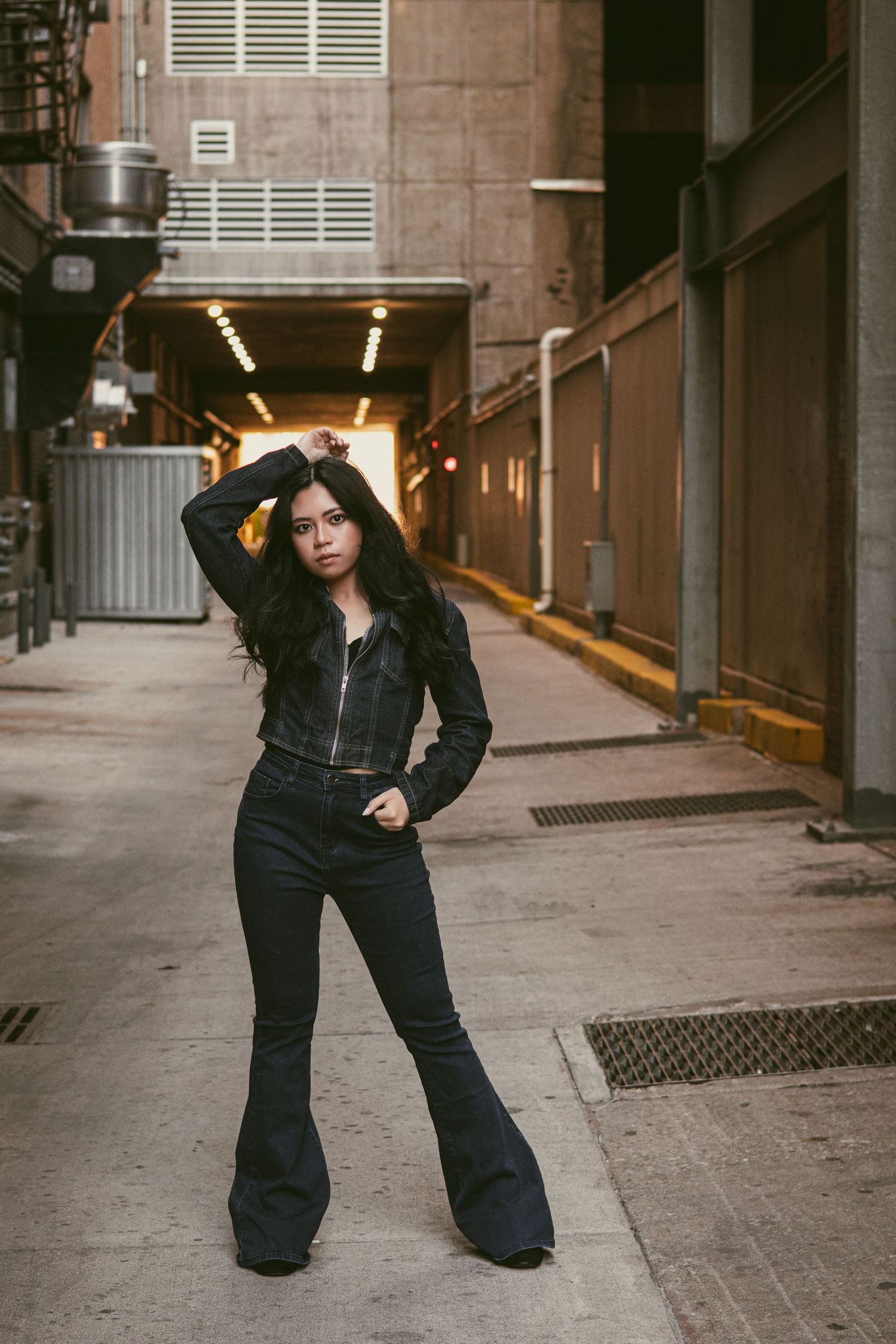 Woman in black jacket and flare jeans poses confidently in city alleyway.