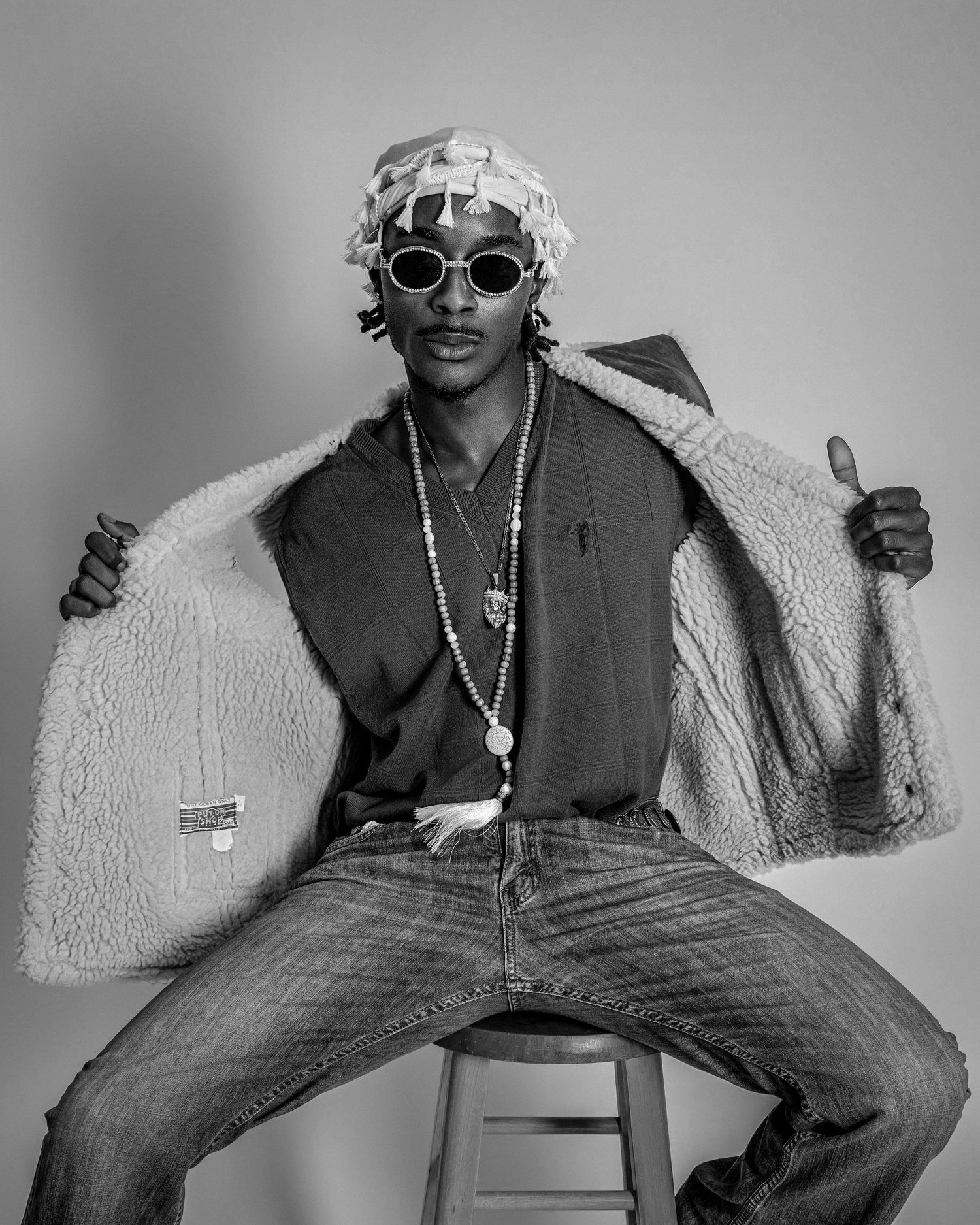 Black and white photo of a stylish man in sunglasses, beanie, sherpa jacket, jeans, and beaded necklace posing on a stool.