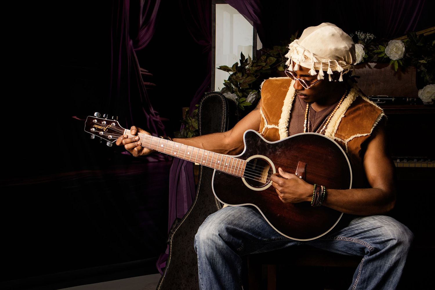 Musician playing acoustic guitar while wearing decorative headpiece, seated against dark backdrop with purple curtains.