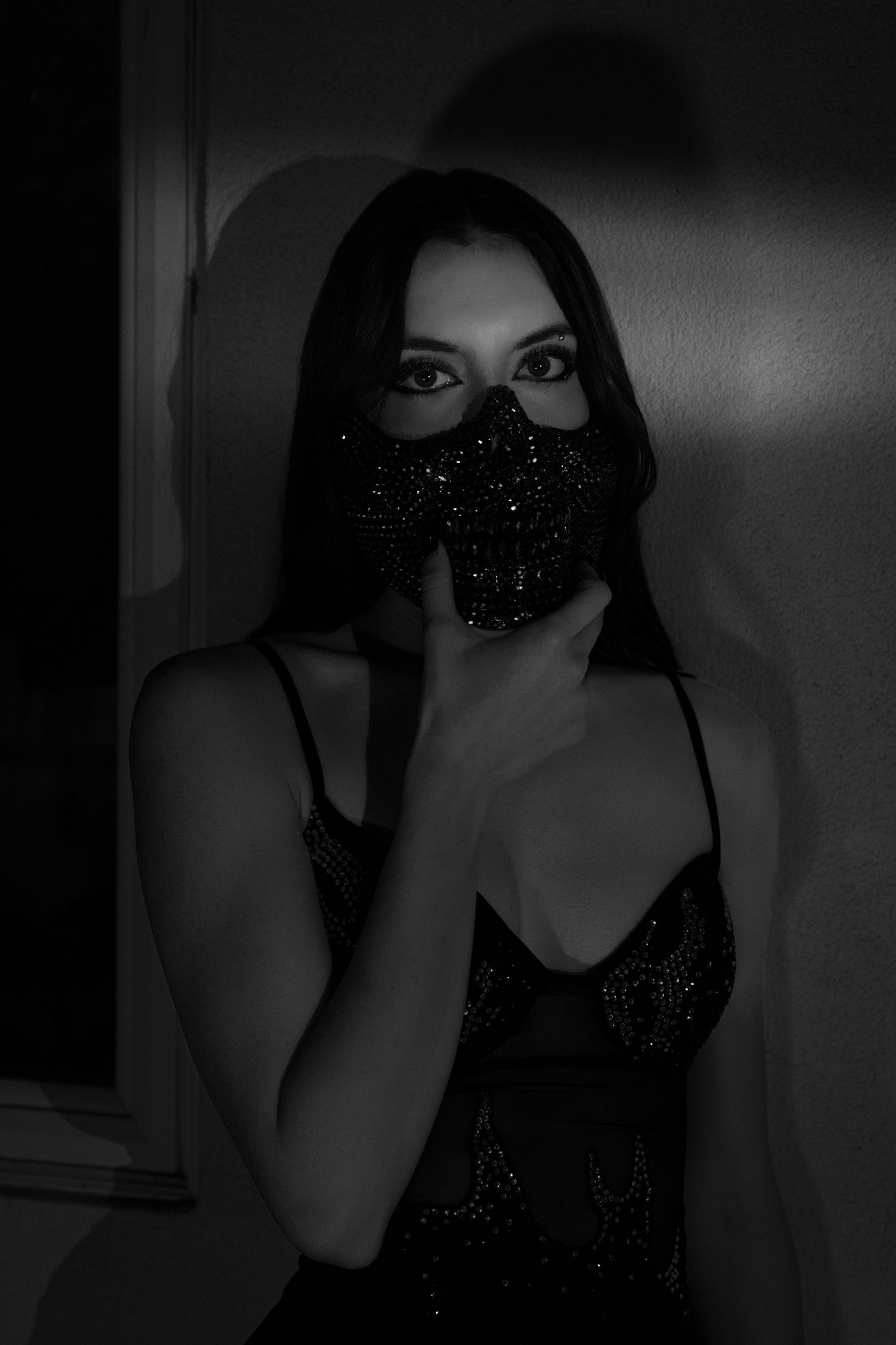 Black and white photo of a woman in a sparkly dress wearing a glittery black face mask, posed against a dark wall.