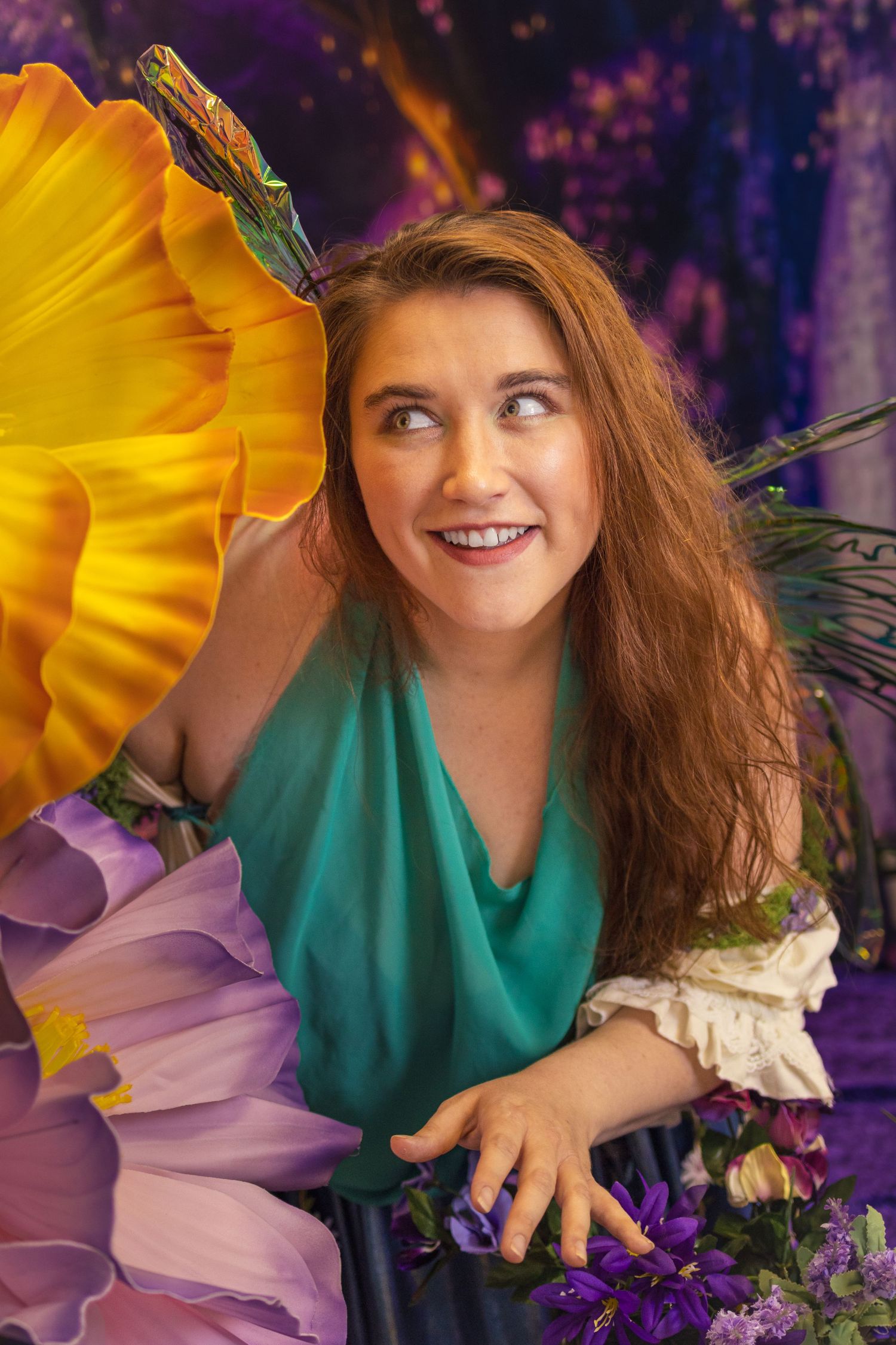 Smiling woman with long red hair in teal dress surrounded by colorful flowers and butterfly wings on purple backdrop.