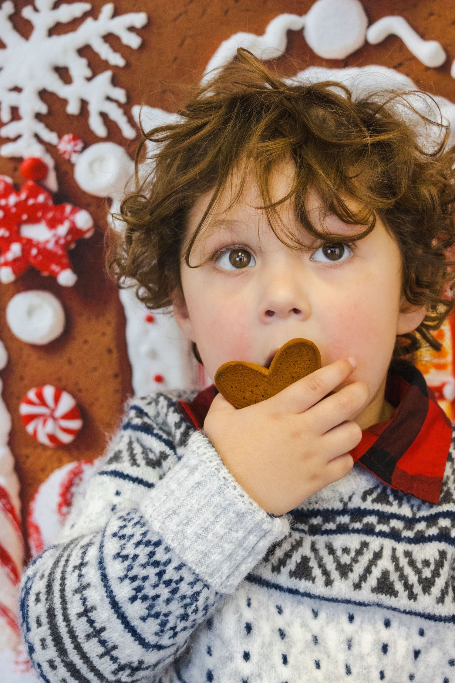 Curly-haired child in Nordic sweater eating a heart-shaped gingerbread cookie near festive Christmas decorations.
