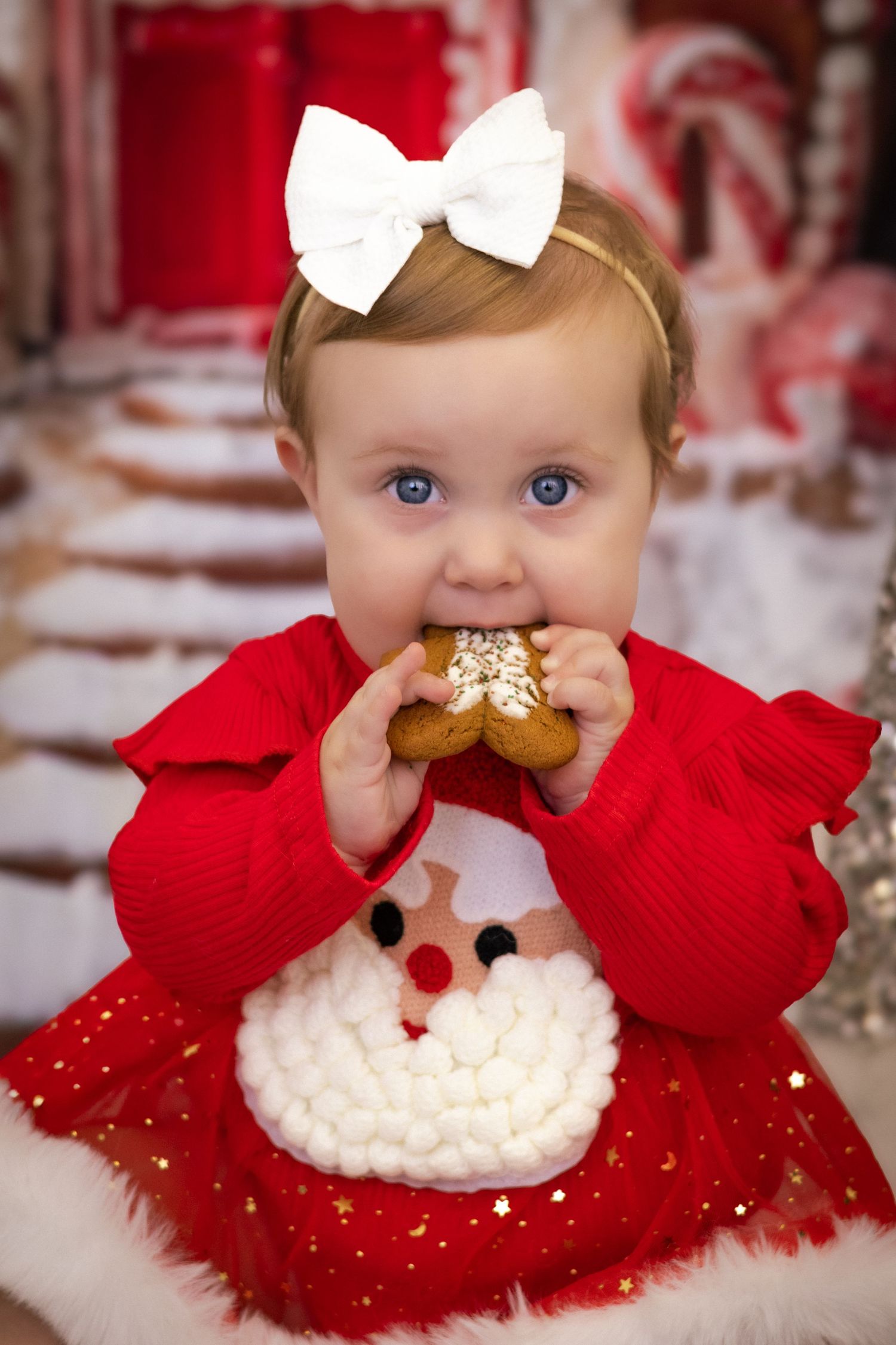 Baby girl in red Santa sweater and white bow eating a Christmas cookie in a festive candy cane decorated setting.
