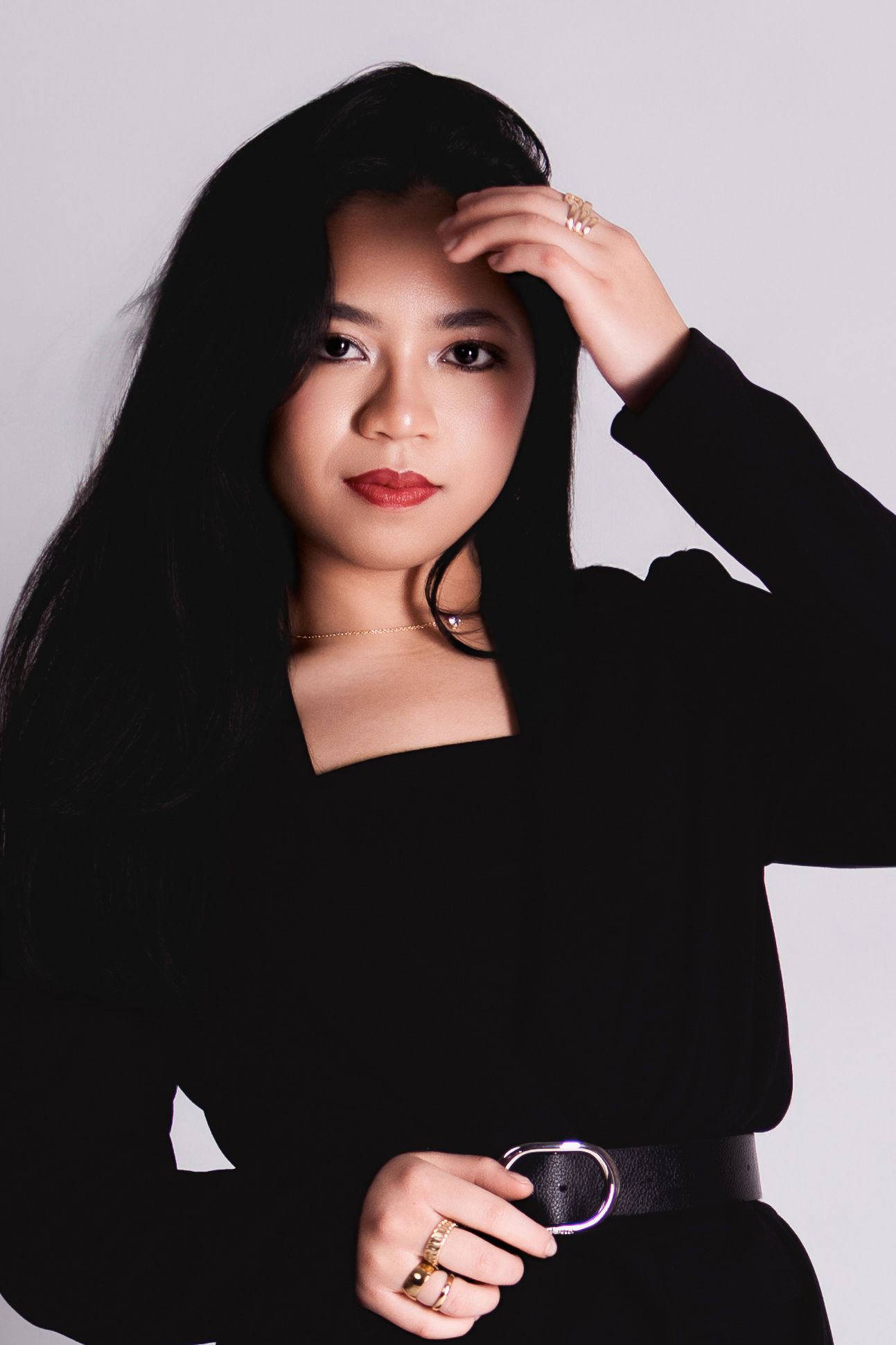 Young Asian woman with long black hair in black top poses confidently against gray background wearing gold rings.
