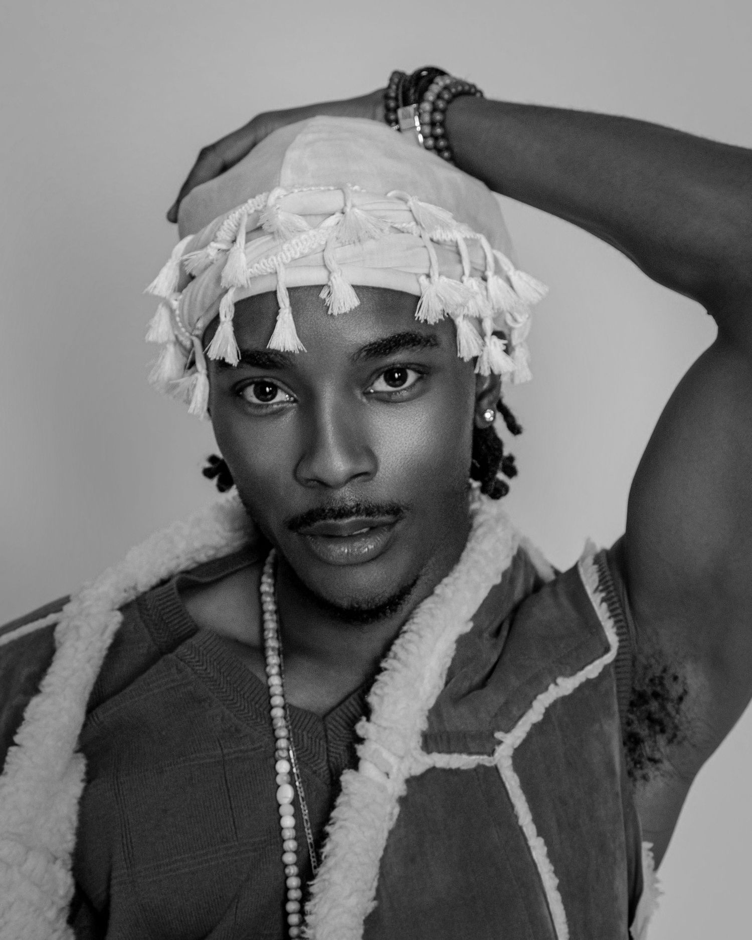 Black and white portrait of a young man wearing a tasseled head wrap and beaded necklace in a stylish editorial pose.