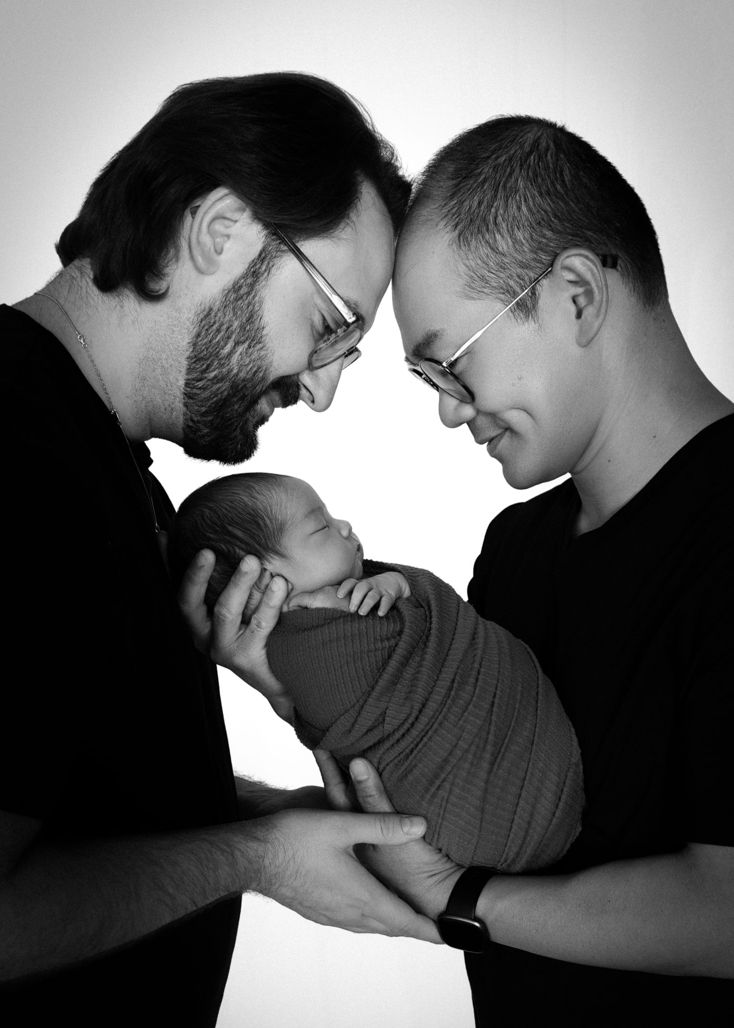Black and white photo of two men with glasses tenderly holding a swaddled newborn baby, touching foreheads together.