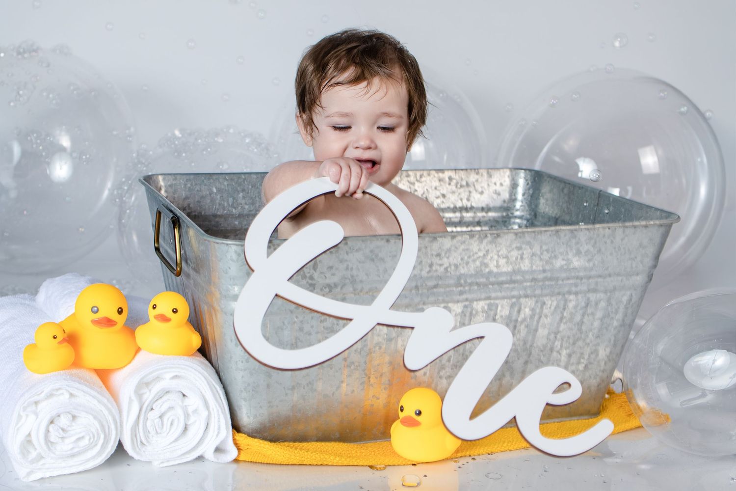 Baby in galvanized tub with rubber ducks and One sign for first birthday.