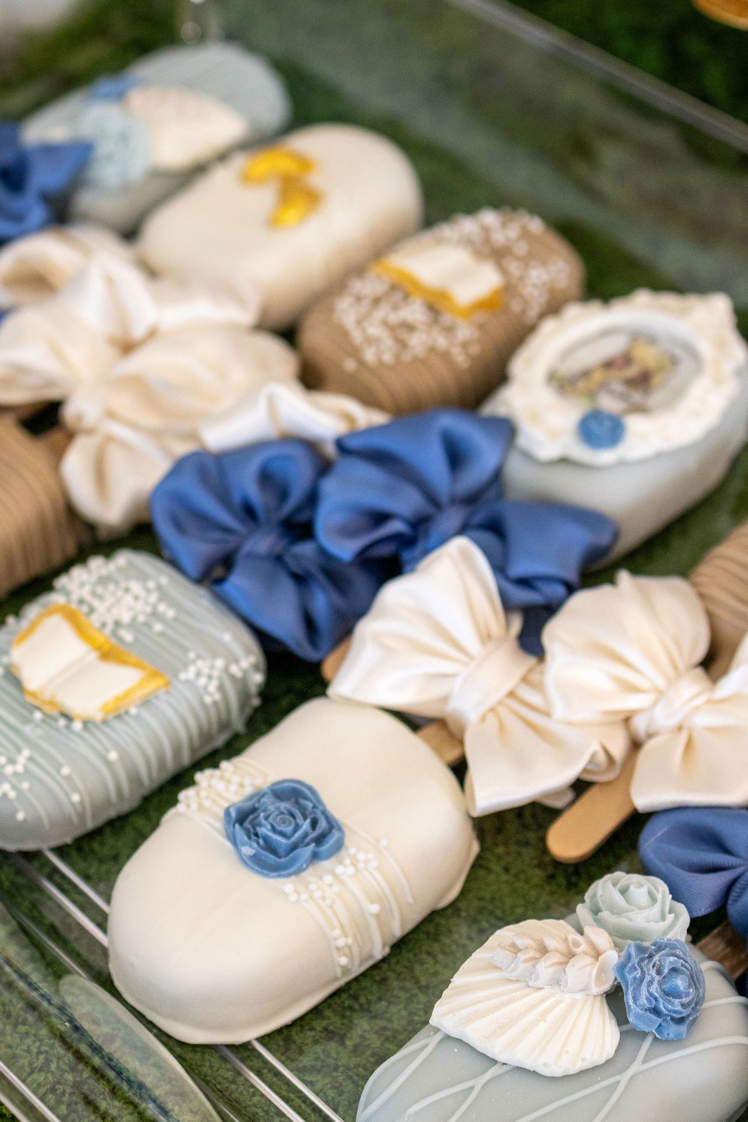 Elegant decorated cake pops with blue and white fondant bows, roses, and pearl details displayed on a tray.