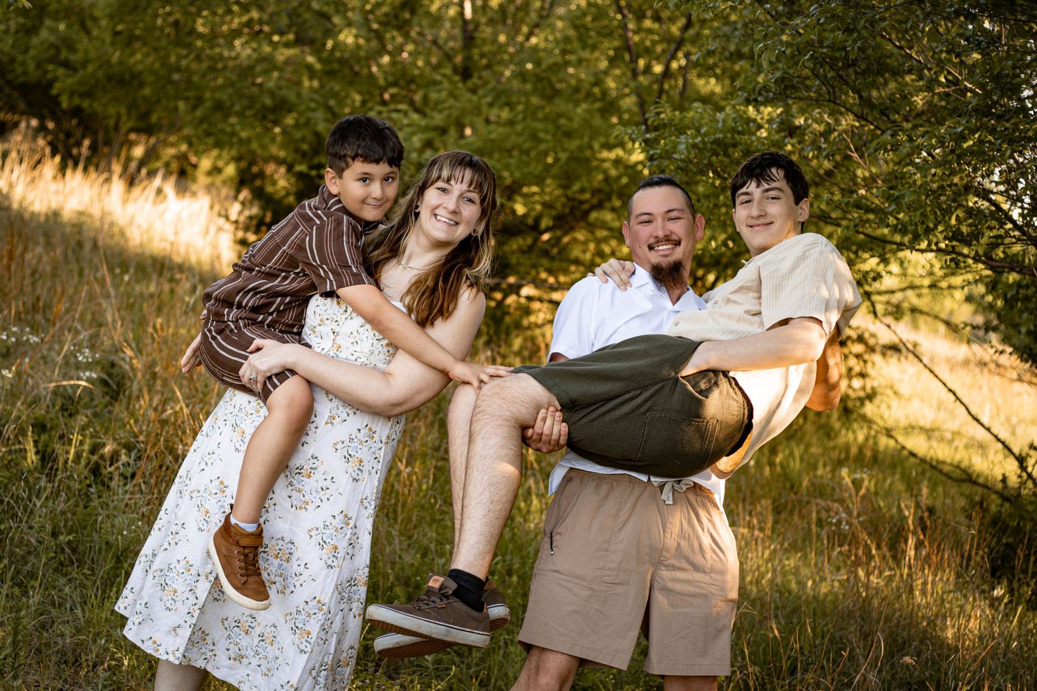 Family of four poses outdoors in a sunny meadow, parents holding two children while smiling near lush green trees.