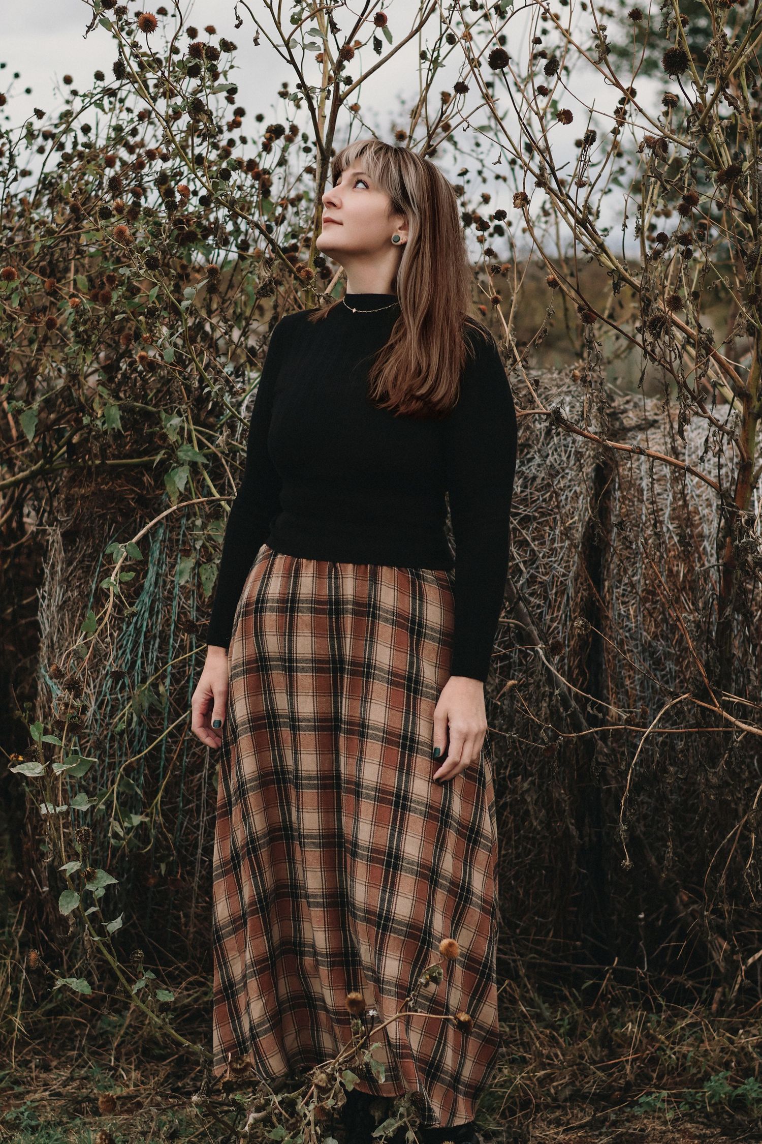 Woman in black turtleneck and plaid maxi skirt standing among autumn branches, gazing upward outdoors.