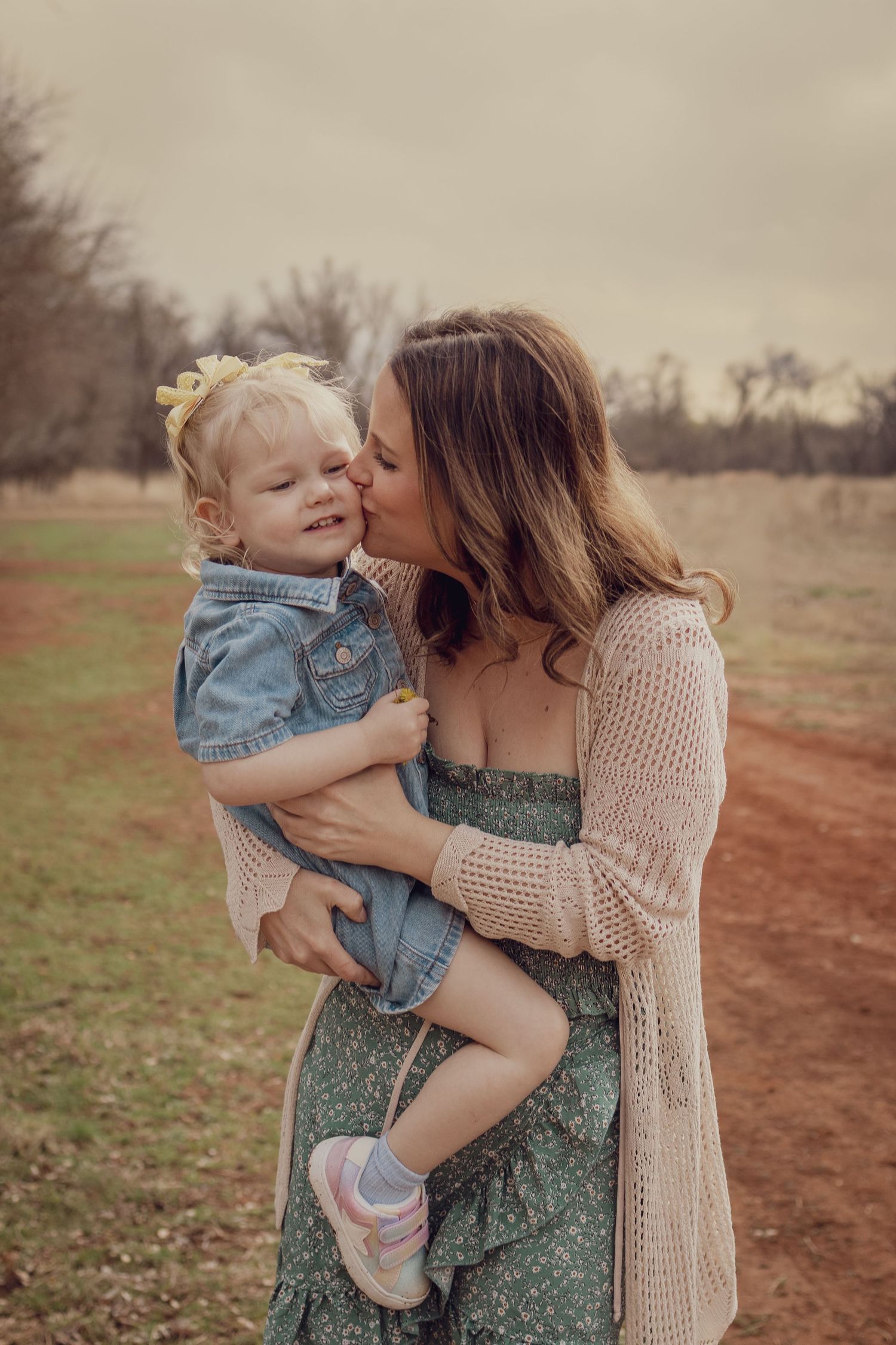 A mother tenderly kisses her smiling toddler girl outdoors, both dressed in casual spring outfits on a dirt path.